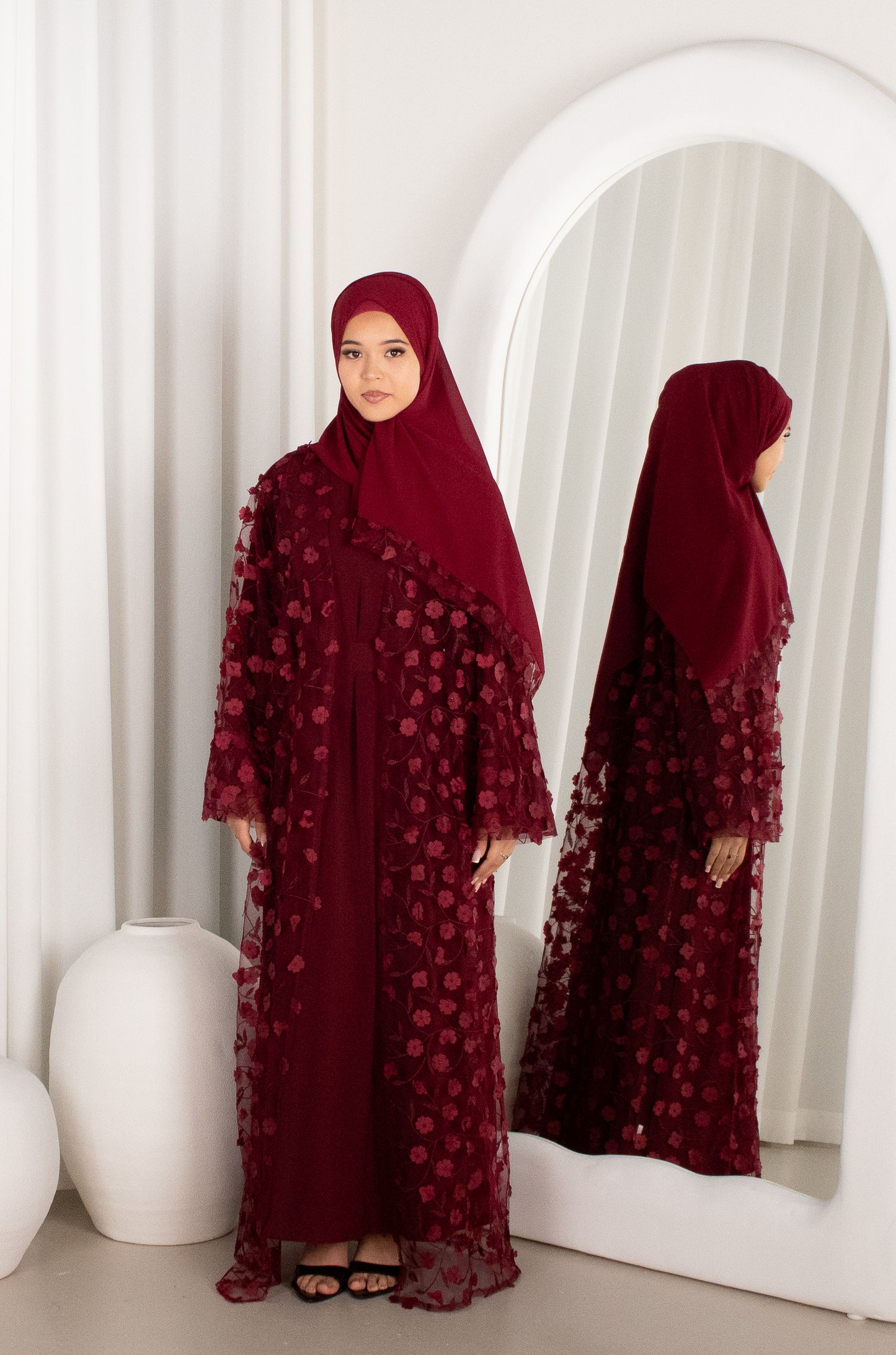 Woman in a burgundy abaya standing in front of a mirror in a white room.