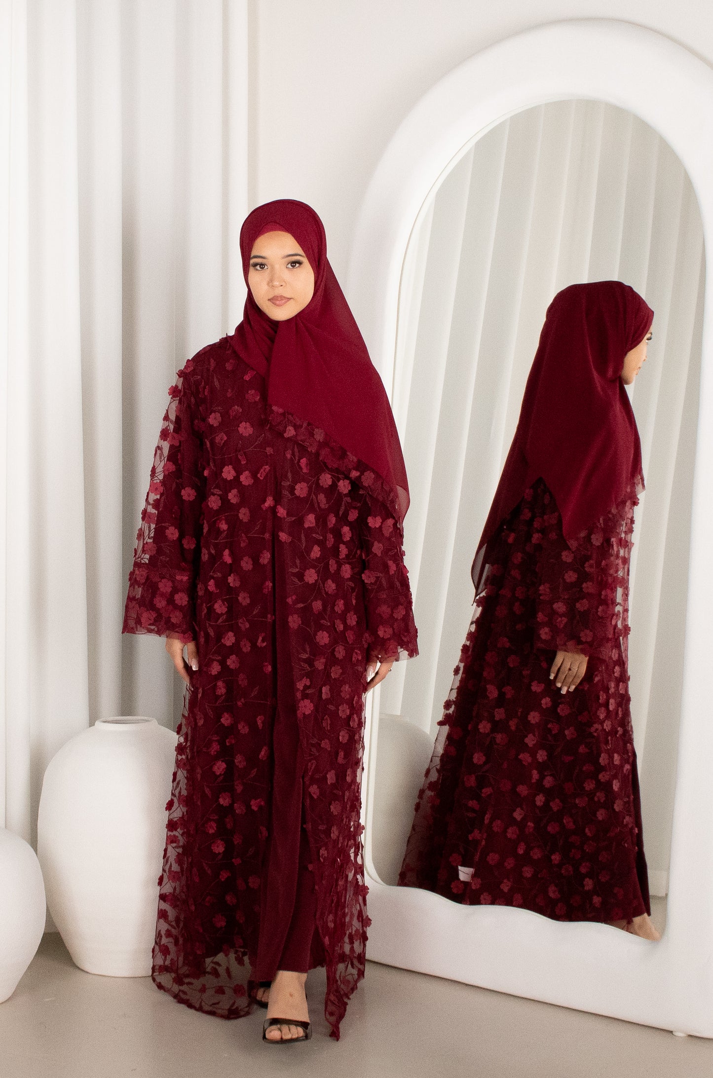 Woman in a burgundy abaya with floral patterns standing in front of a mirror.