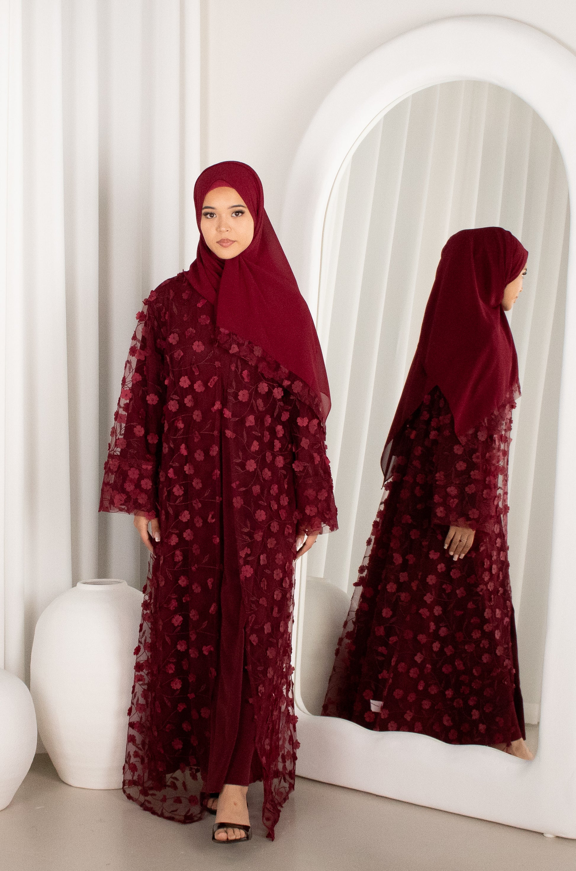 Woman in a burgundy abaya with floral patterns standing in front of a mirror.