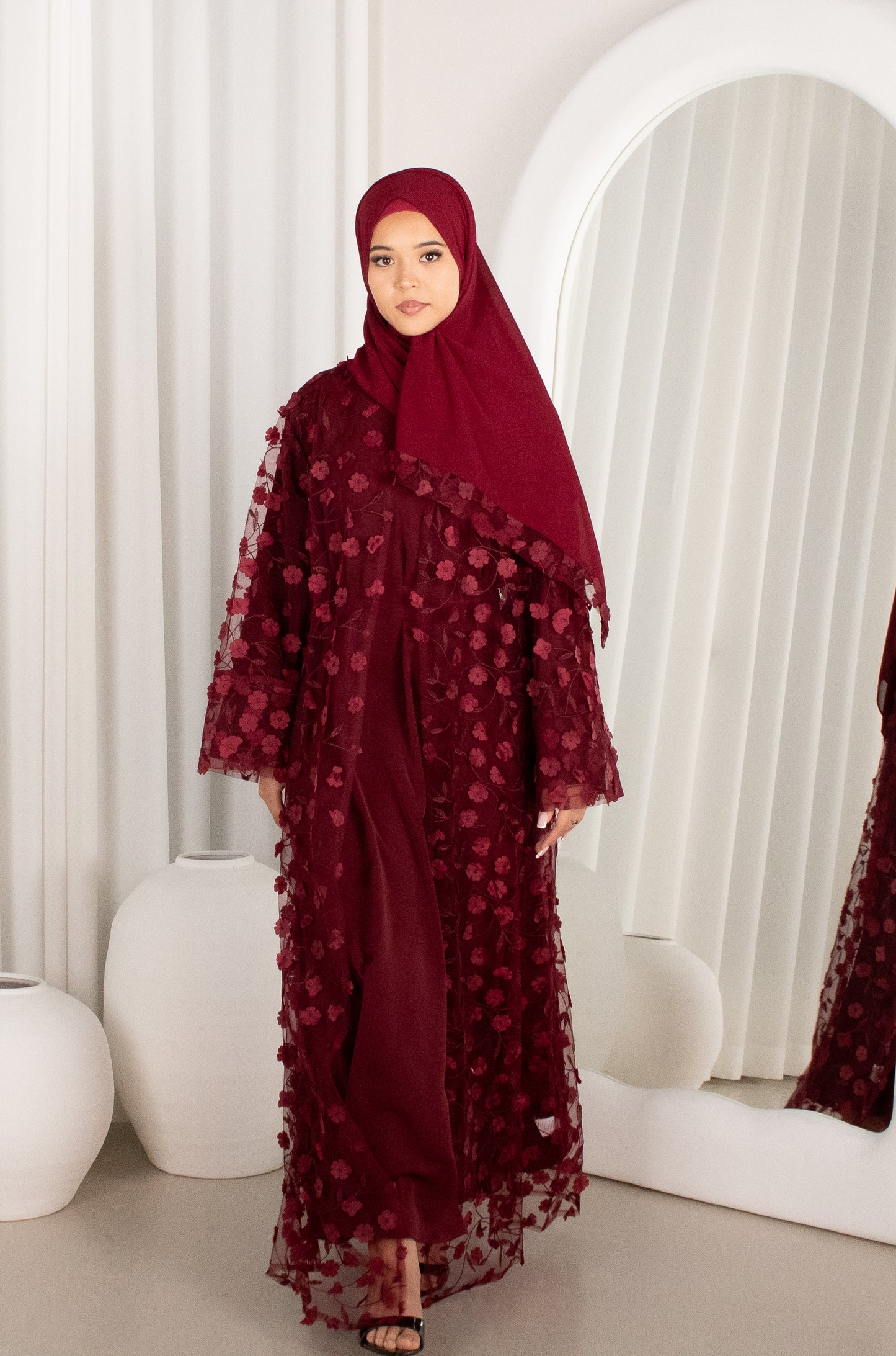 Woman wearing a burgundy floral dress and hijab in a minimalistic room.