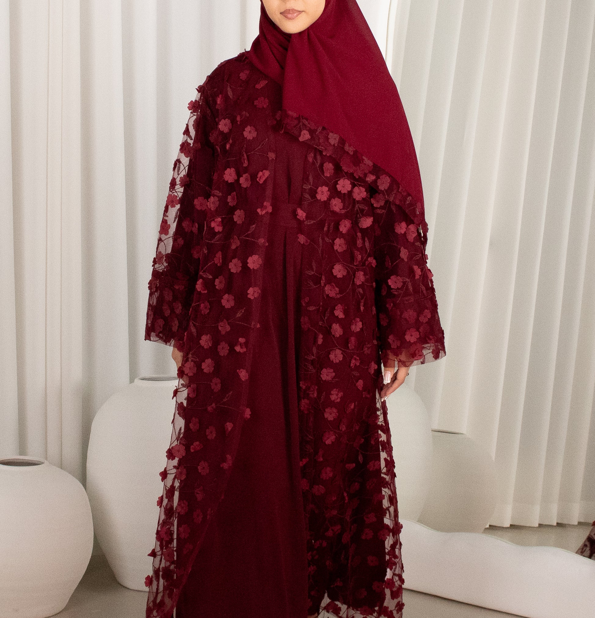 Person wearing a burgundy abaya with floral embroidery against a white curtain background