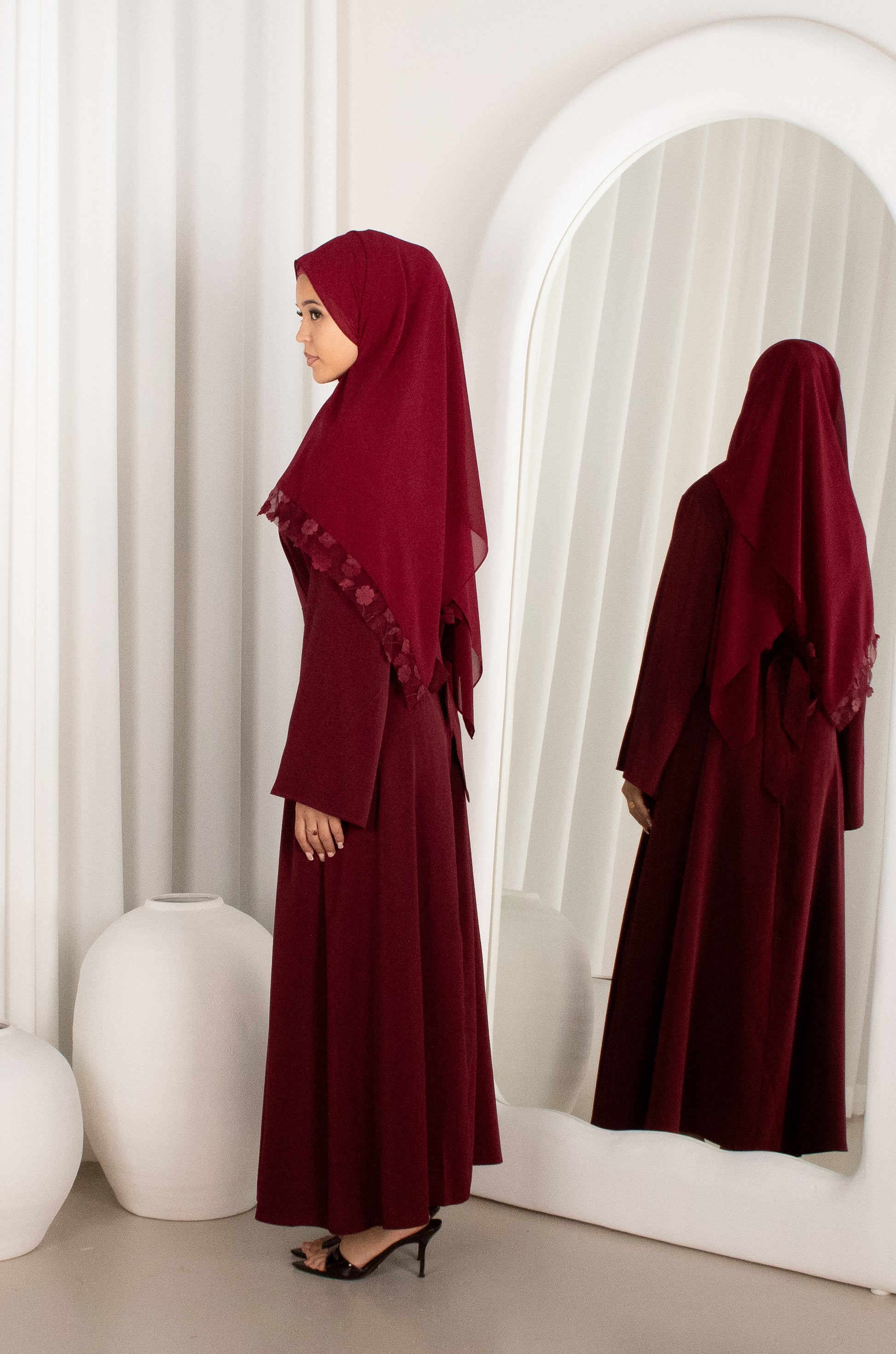Woman in a burgundy abaya standing in front of a mirror in a minimalistic setting.