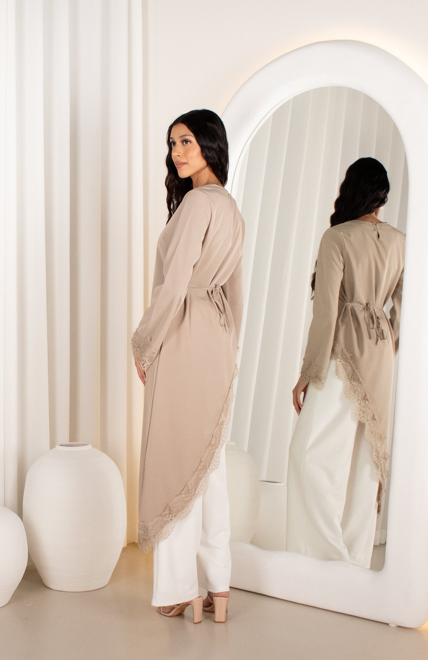 Woman in a beige cardigan and white pants standing in front of a mirror in a minimalistic room.