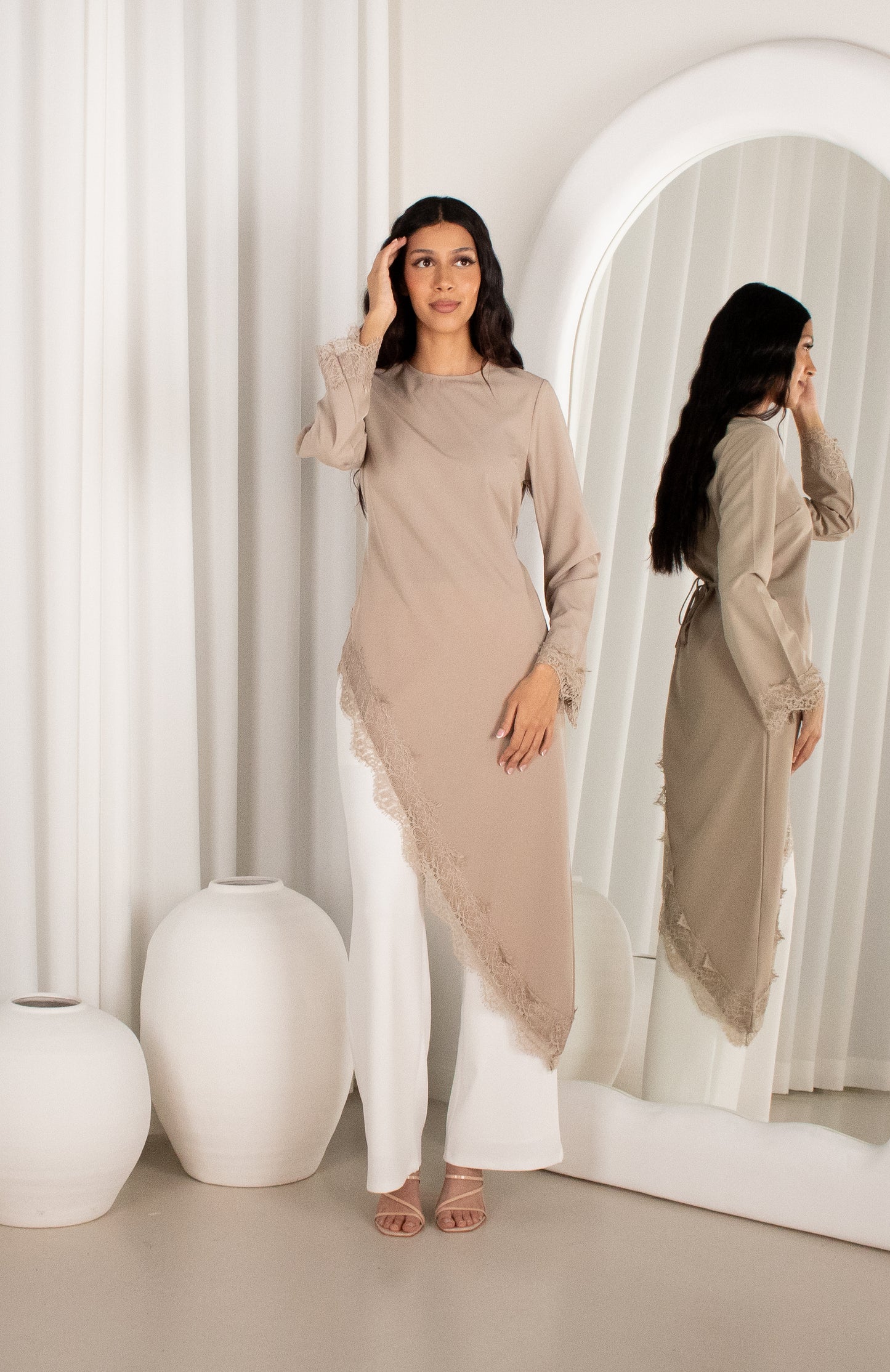 Woman in a beige long-sleeve top and white pants standing in front of a mirror.