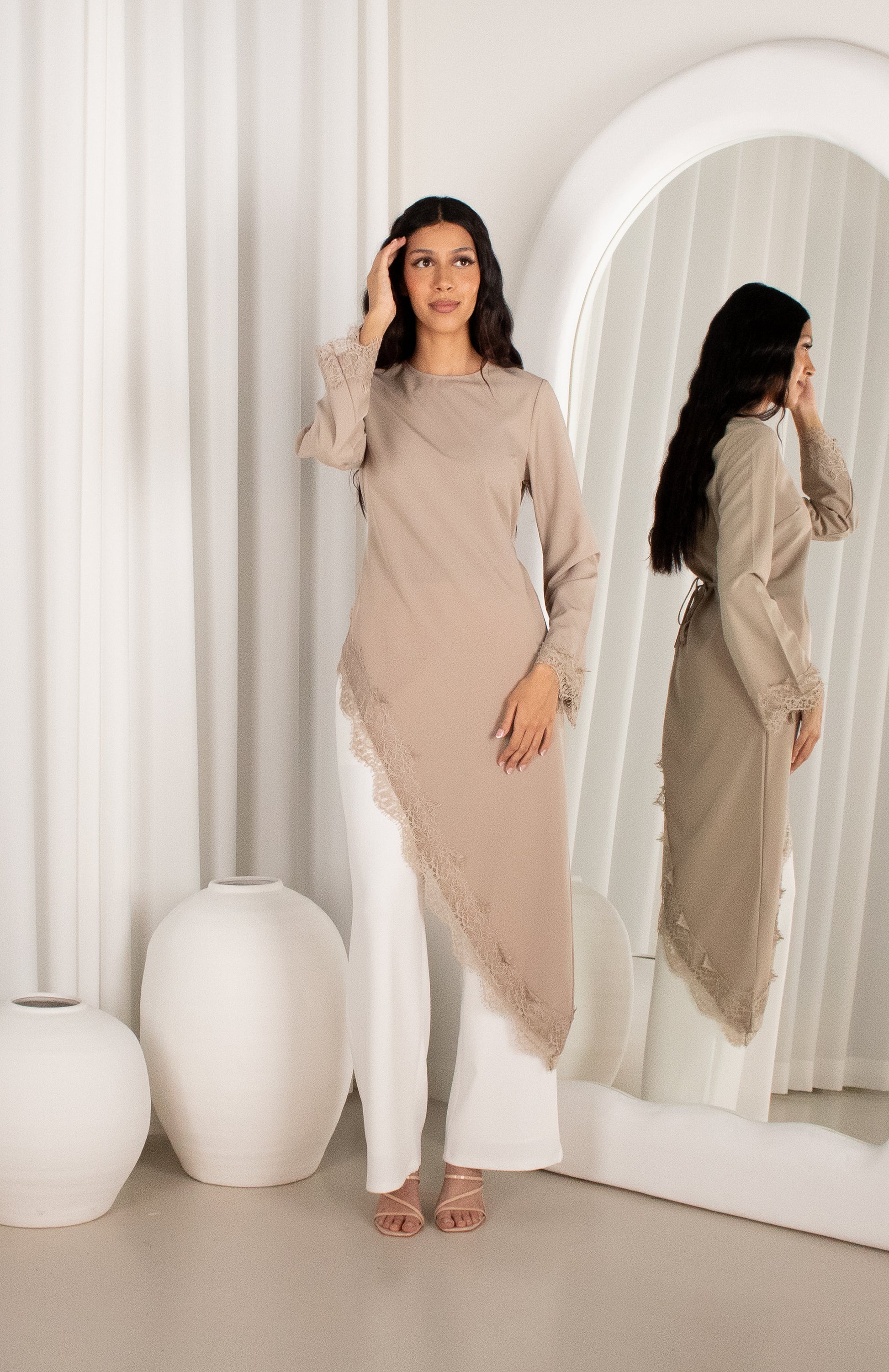 Woman in a beige long-sleeve top and white pants standing in front of a mirror.