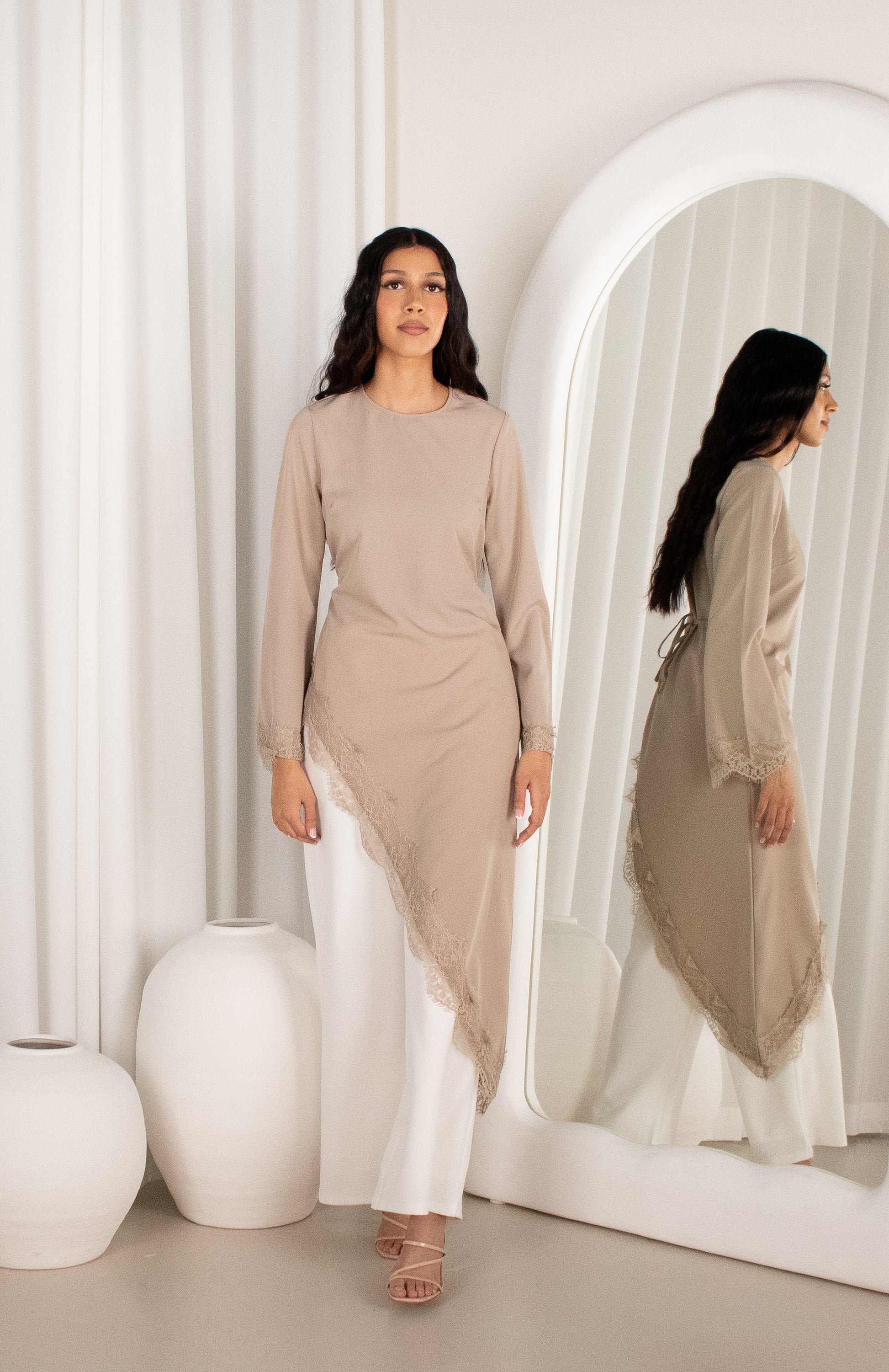 Woman in a beige top and white pants standing in front of a mirror in a minimalistic room.