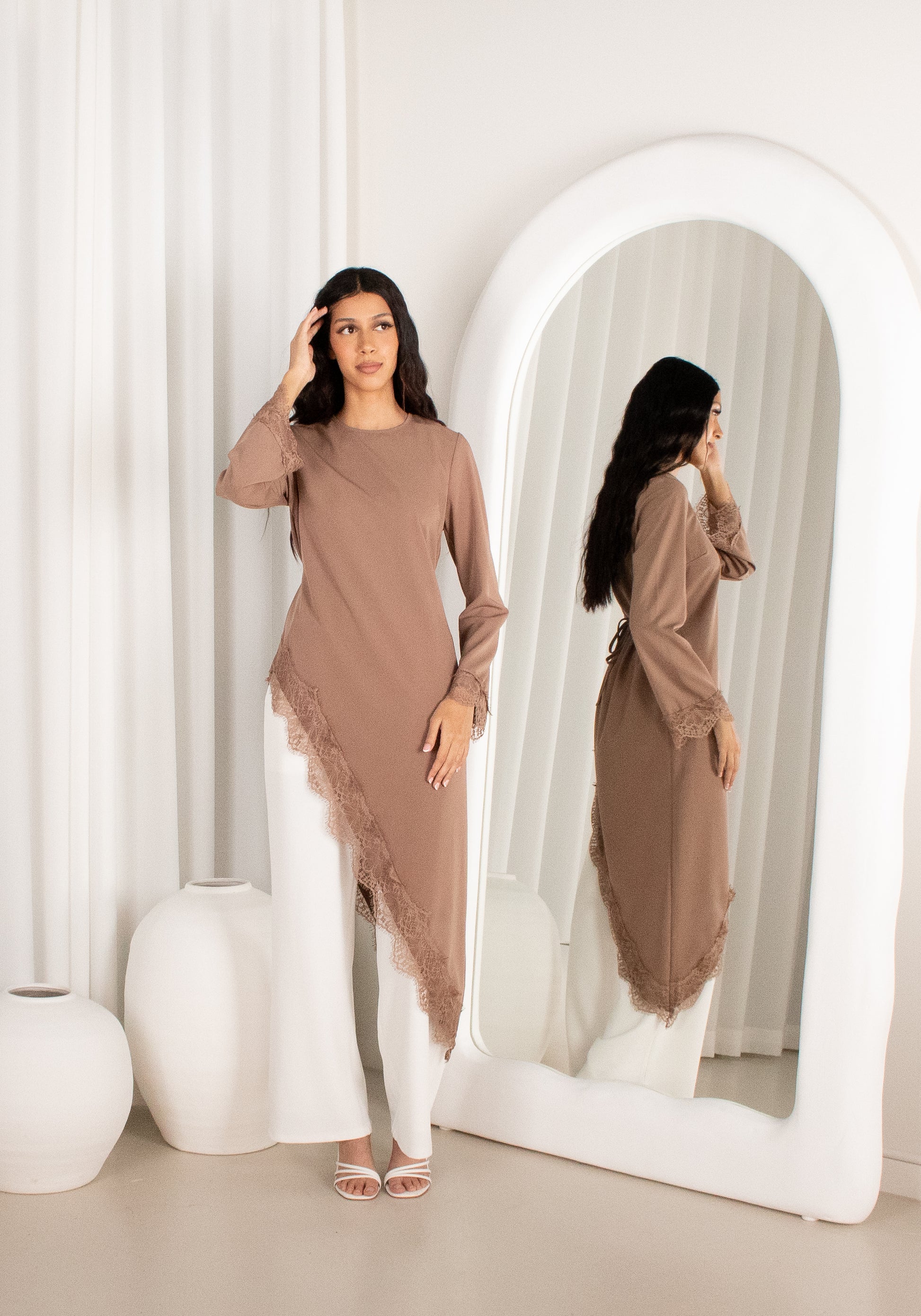 Woman in a brown outfit standing in front of a mirror in a minimalistic room.