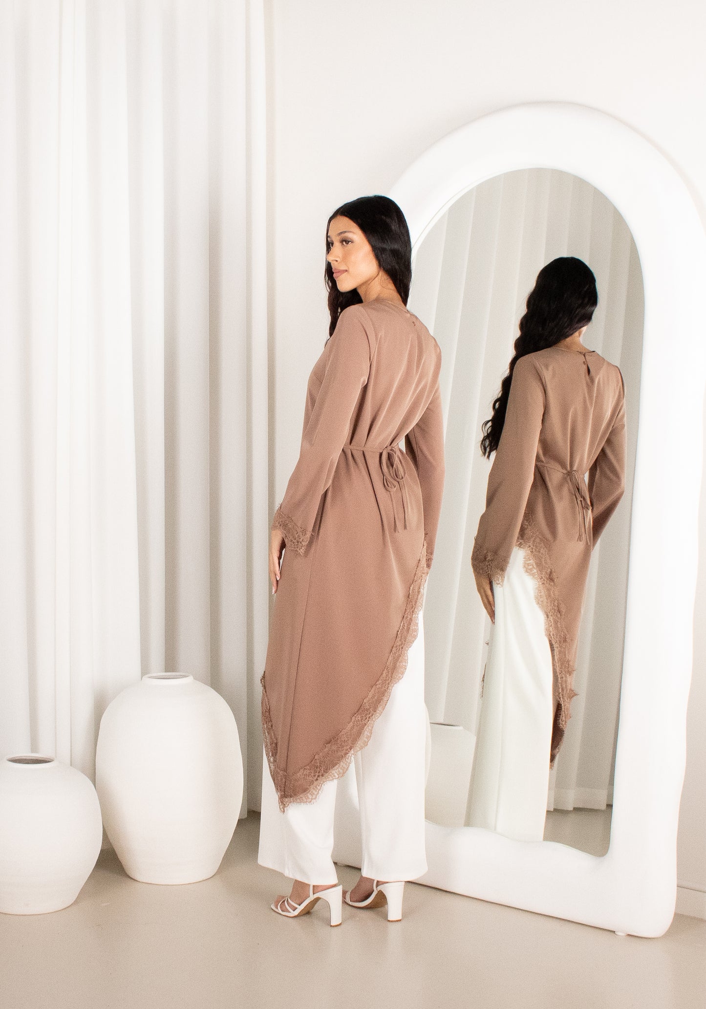 Woman in a long brown cardigan and white pants standing in front of a mirror.