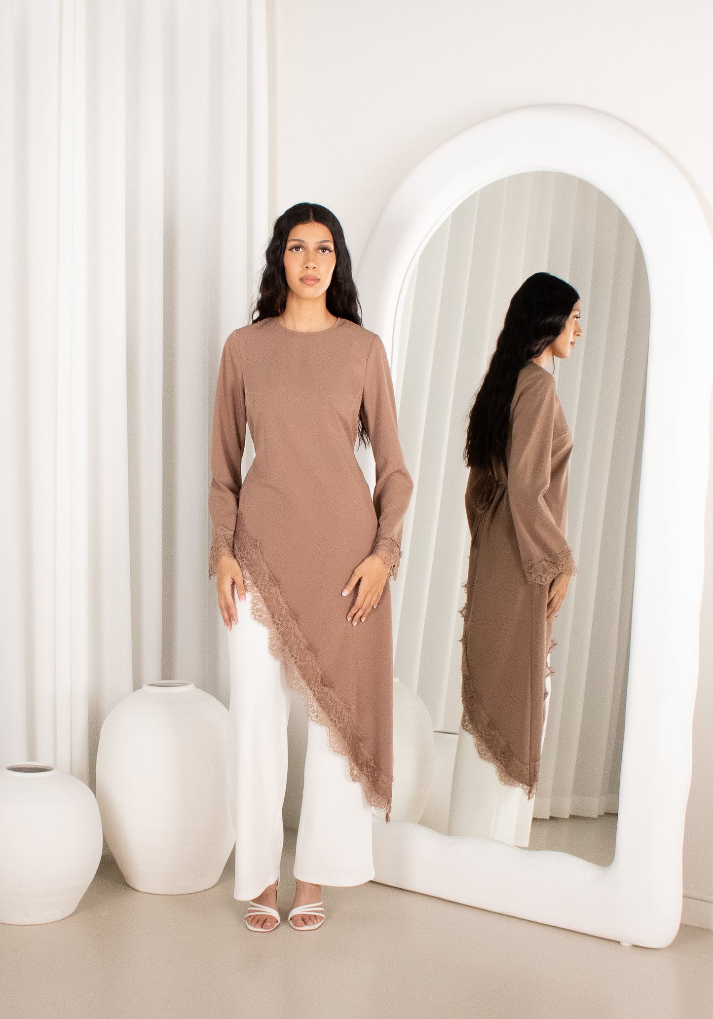 Woman in a brown long-sleeve top and white pants standing in front of a mirror.