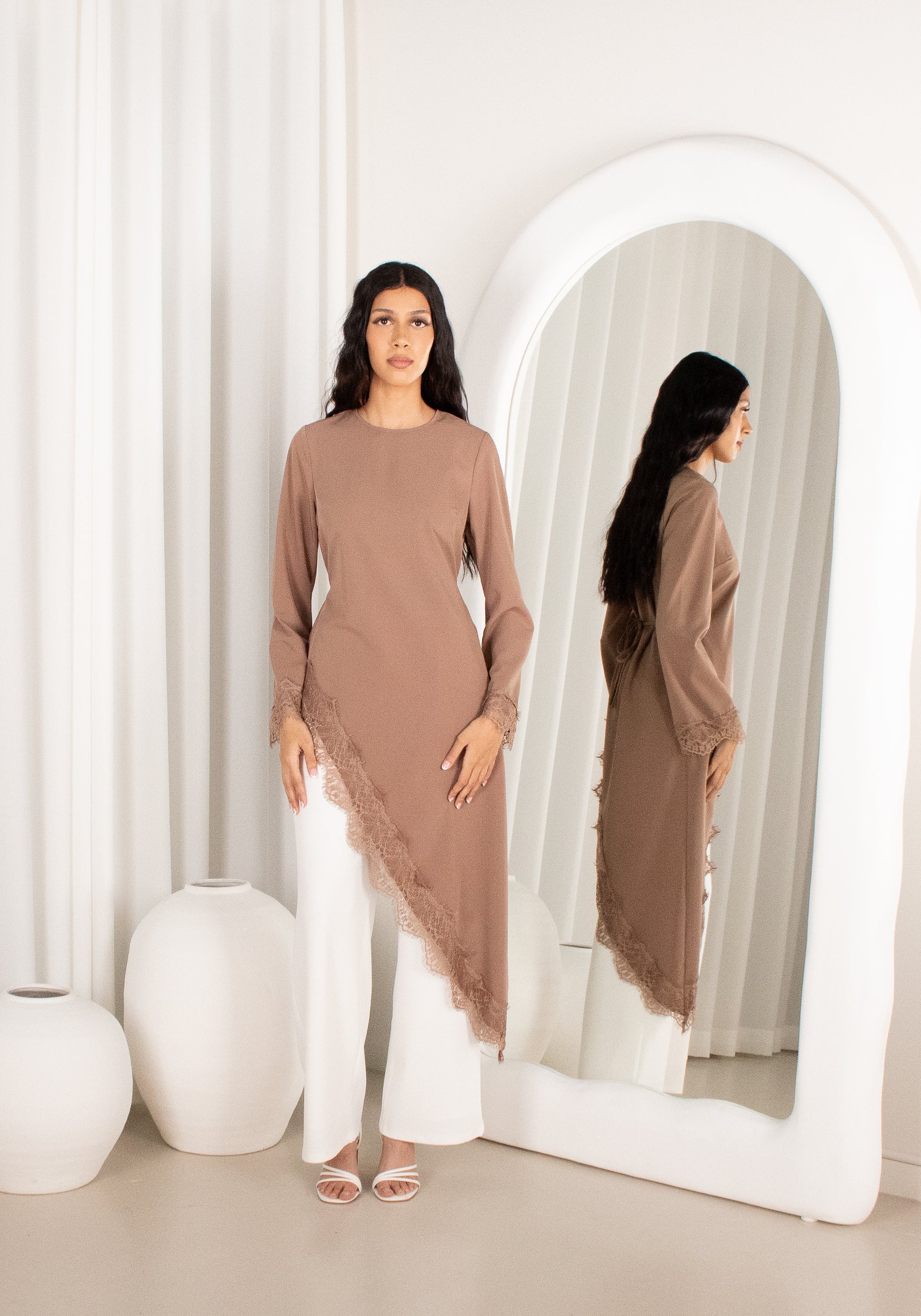Woman in a brown long-sleeve top and white pants standing in front of a mirror.