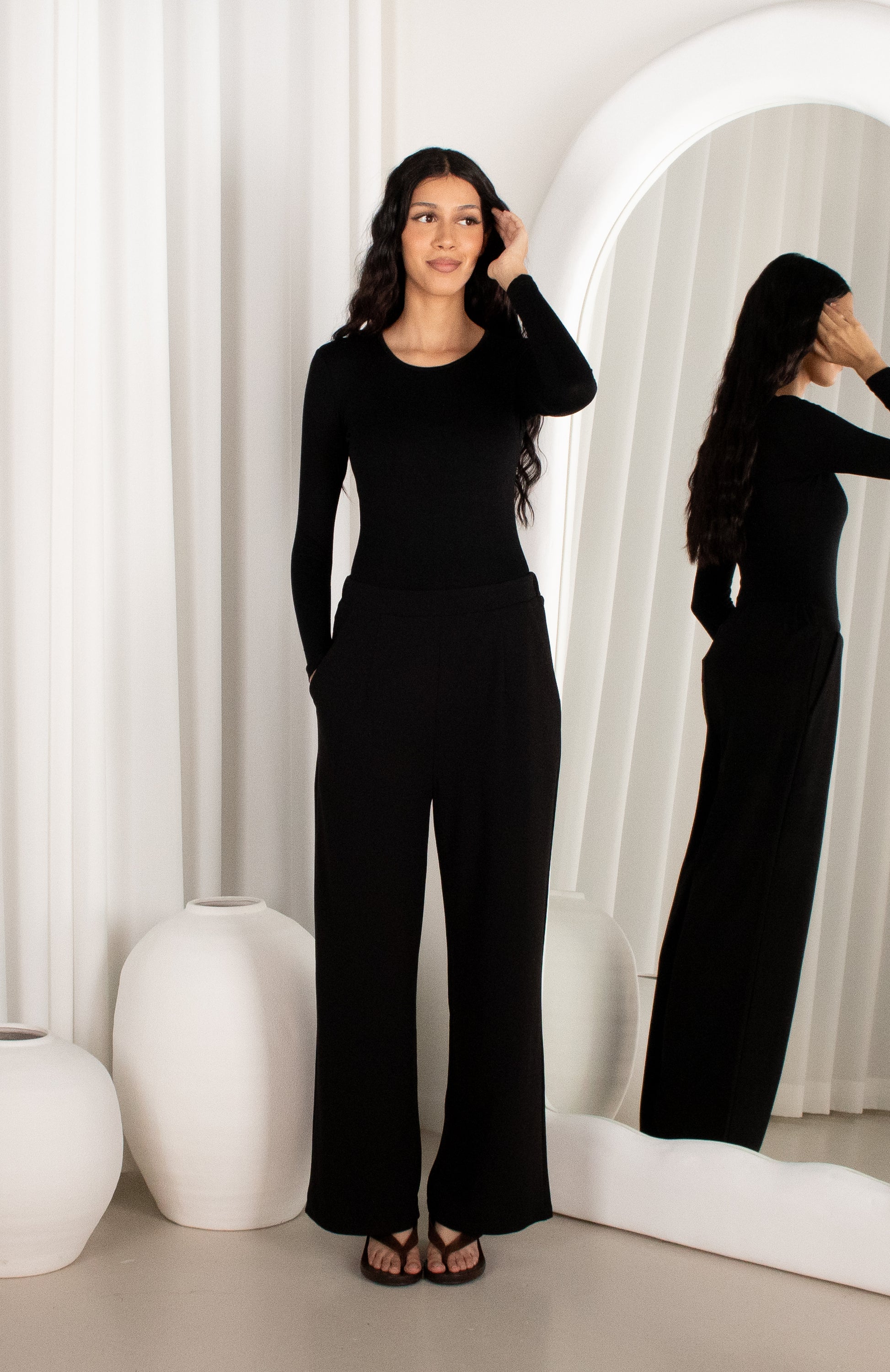 Woman in a black outfit standing in front of a mirror in a minimalistic room.