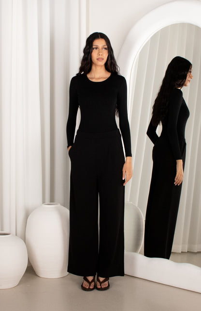Woman wearing a black jumpsuit standing in front of a mirror with white curtains.