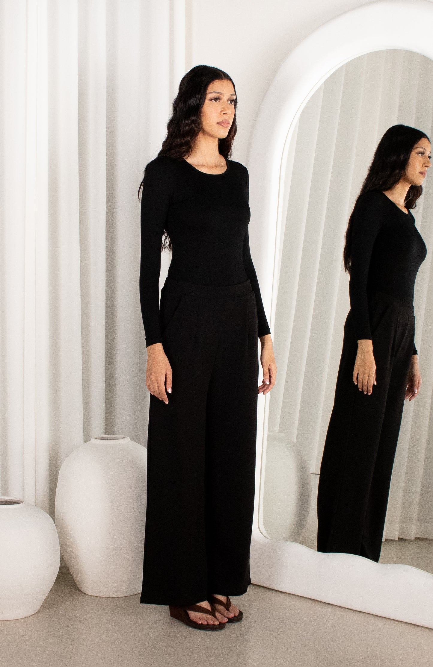 Woman in a black outfit standing in front of a mirror in a minimalistic room.