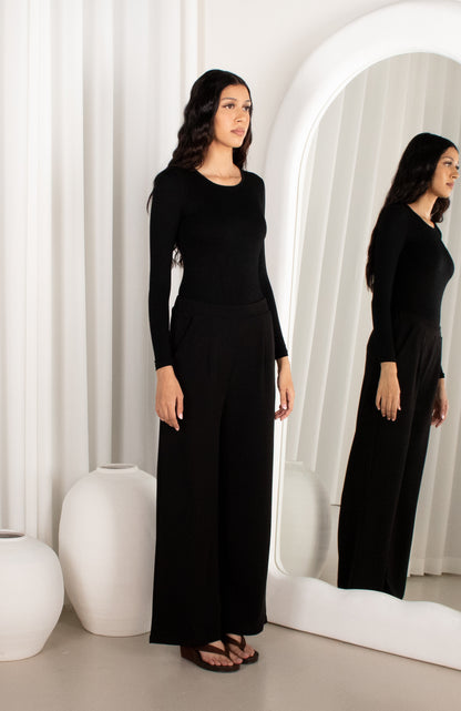 Woman in a black outfit standing in front of a mirror in a minimalistic room.
