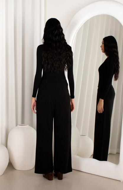 Woman wearing a black jumpsuit standing in front of a mirror.