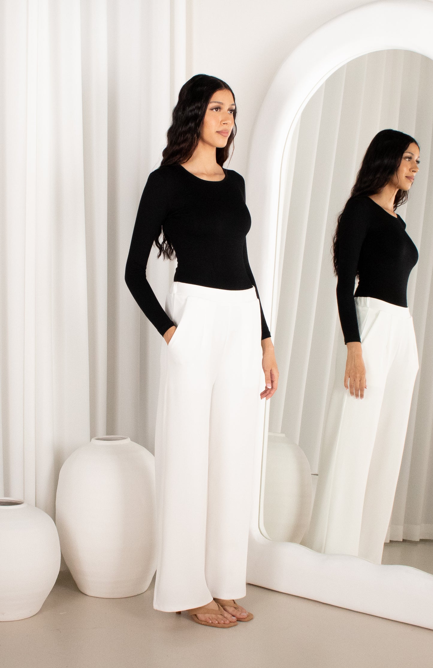 Woman in black top and white pants standing in front of a mirror with white curtains.