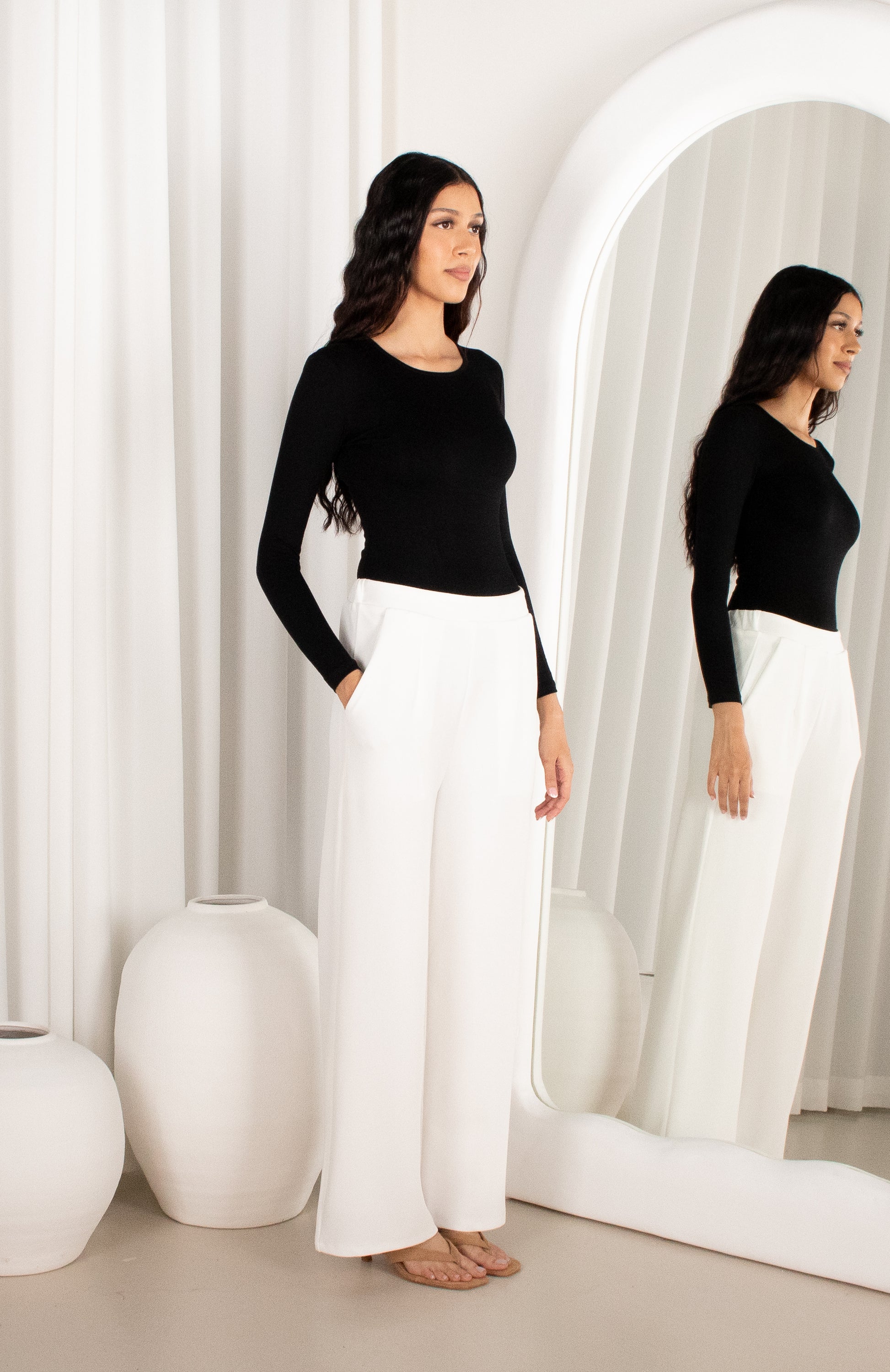 Woman in black top and white pants standing in front of a mirror with white curtains.