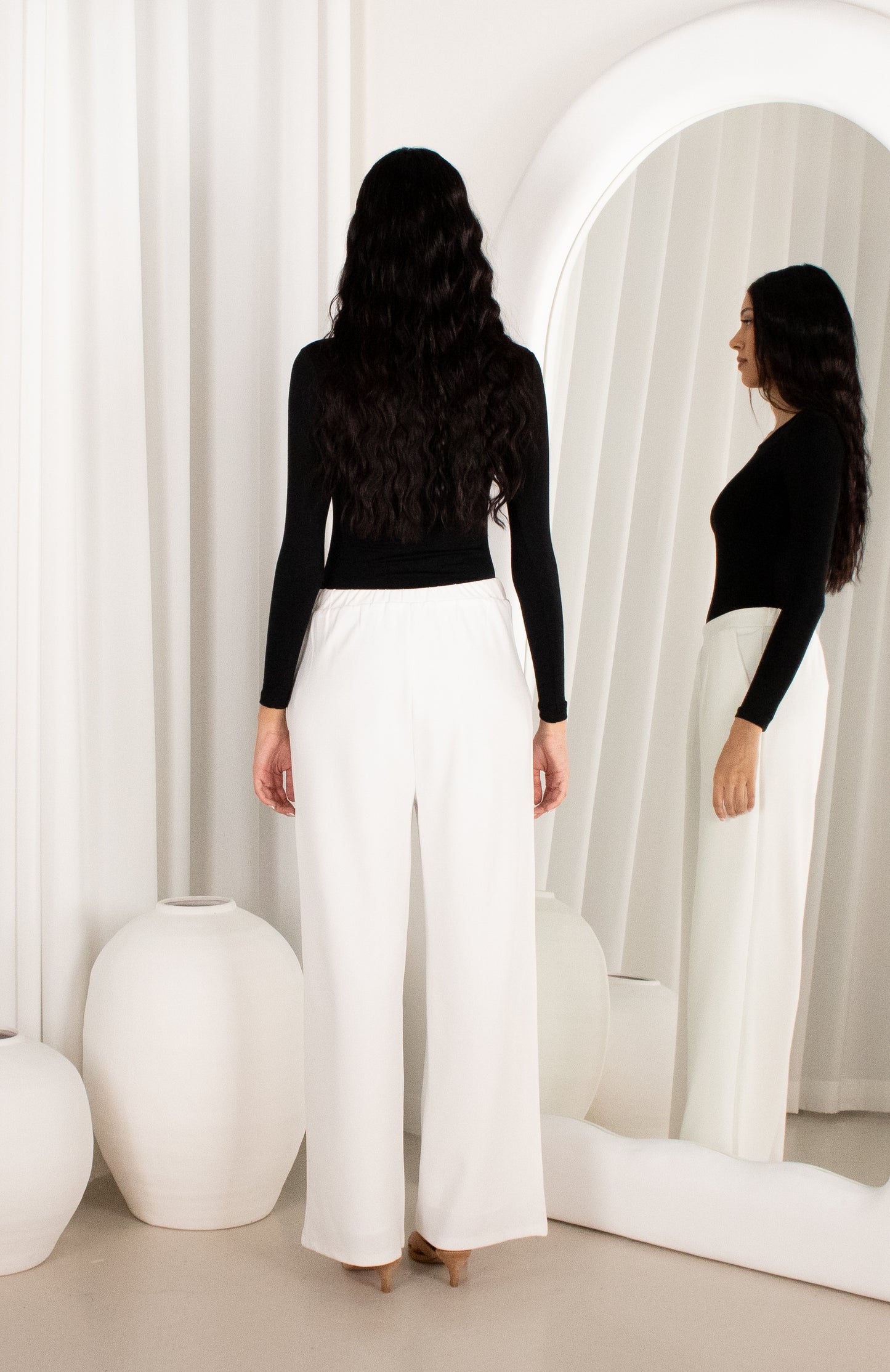 Woman wearing a black top and white pants standing in front of a mirror.