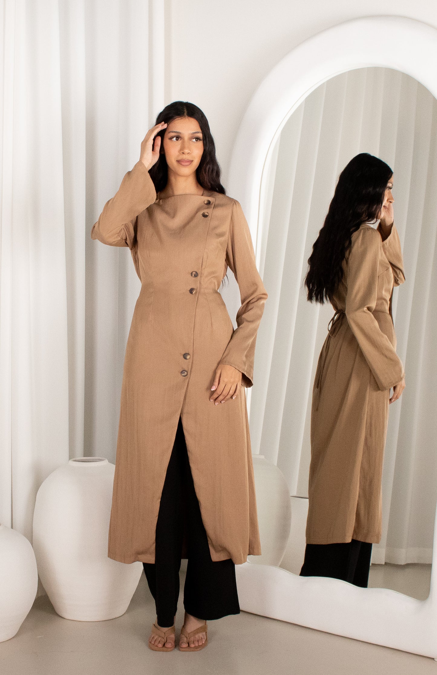 Woman wearing a long beige coat standing in front of a mirror.