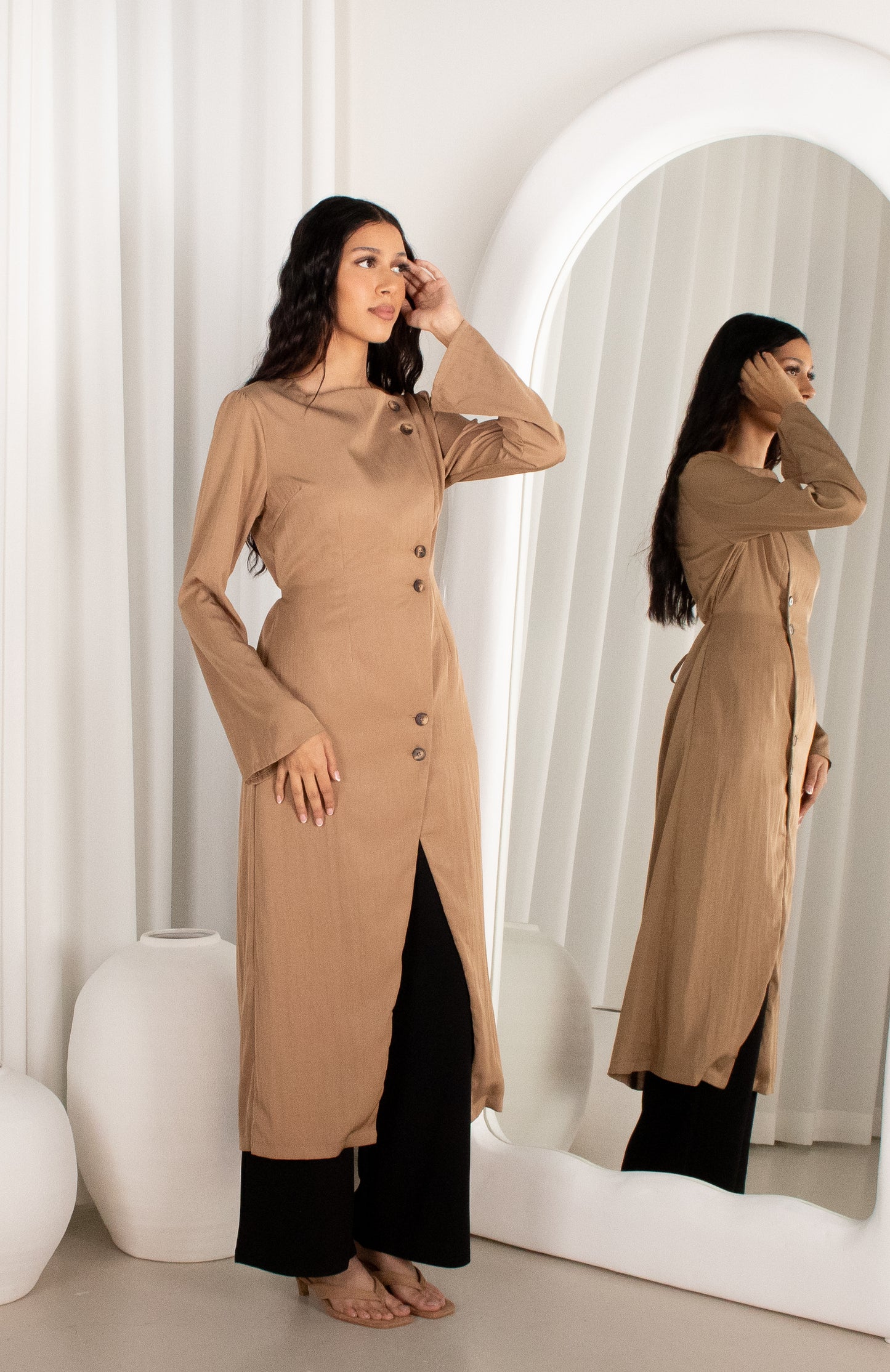 Woman wearing a long beige coat standing in front of a mirror.