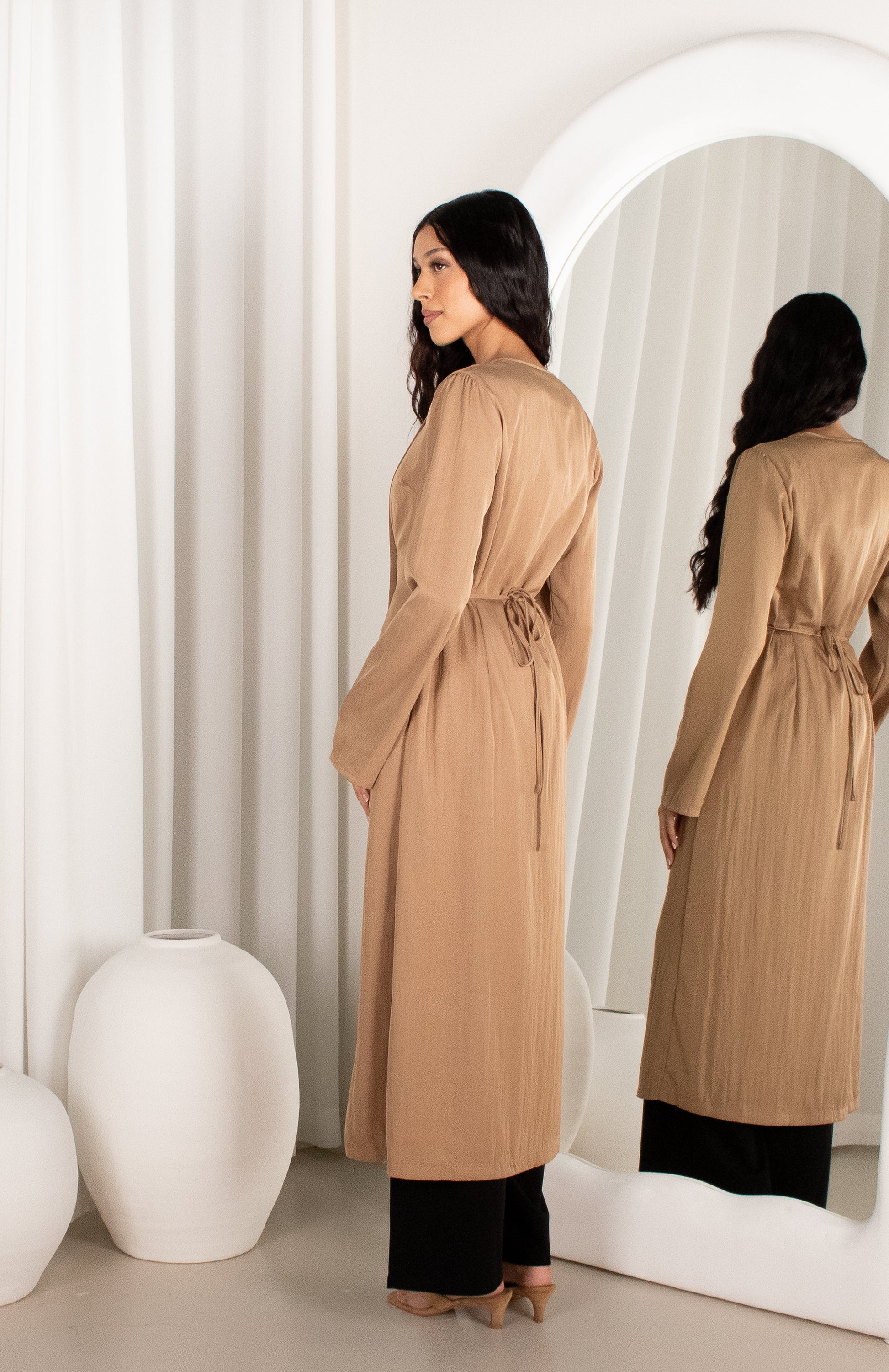 Woman in a beige trench coat standing in front of a mirror in a minimalistic room.