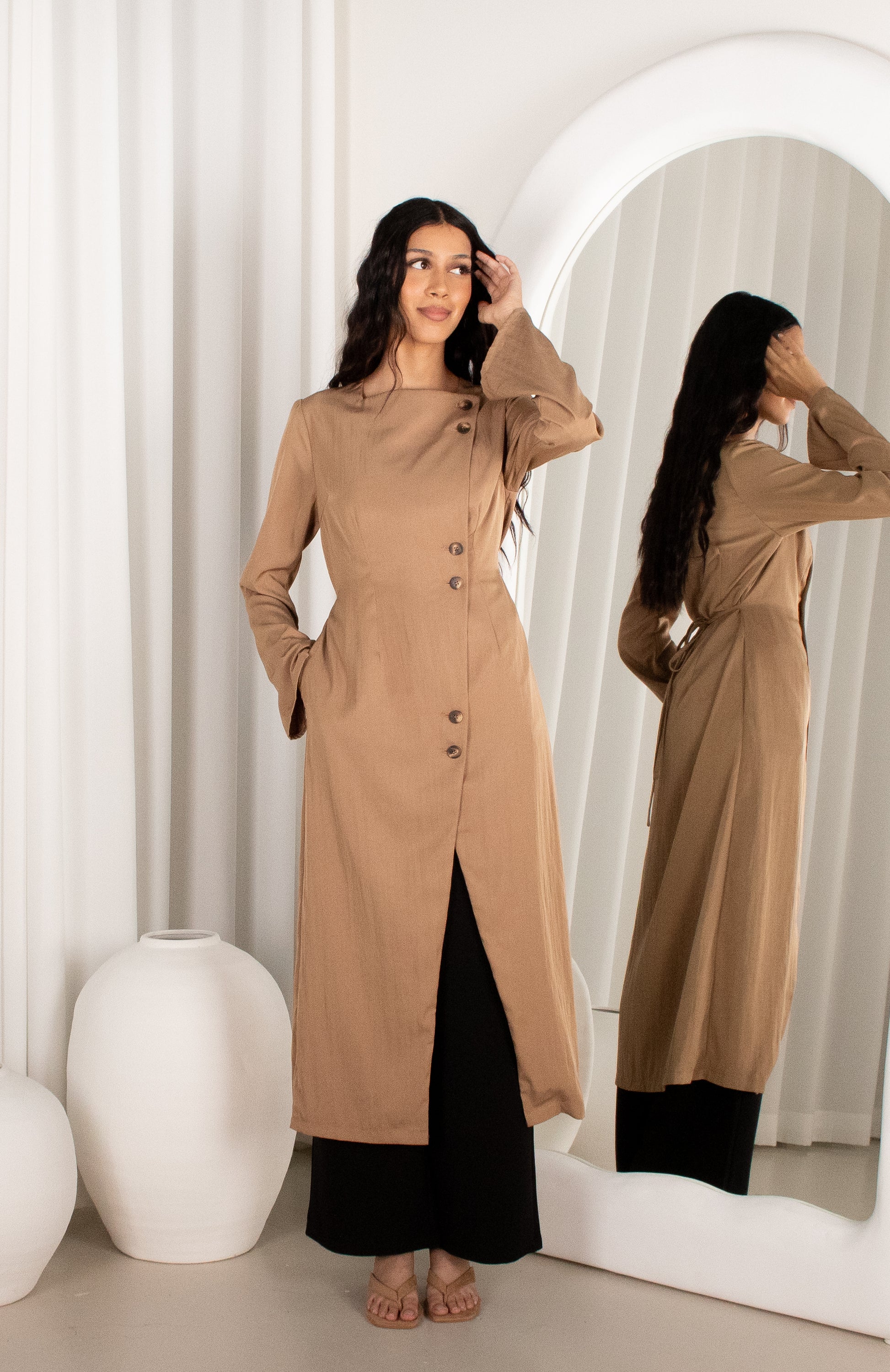 Woman wearing a long beige coat standing in front of a mirror.