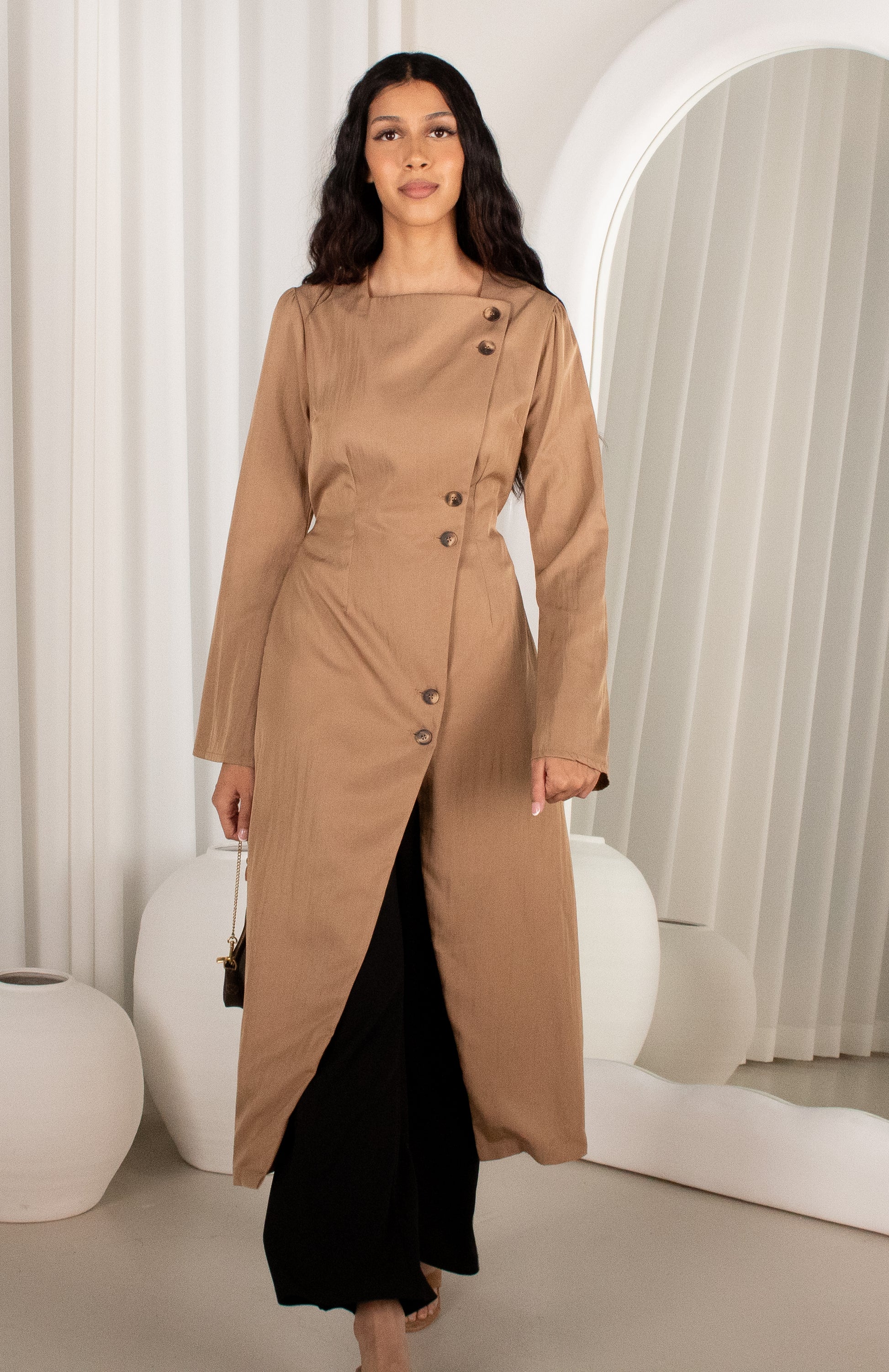 Woman wearing a long beige coat standing in a modern interior setting.