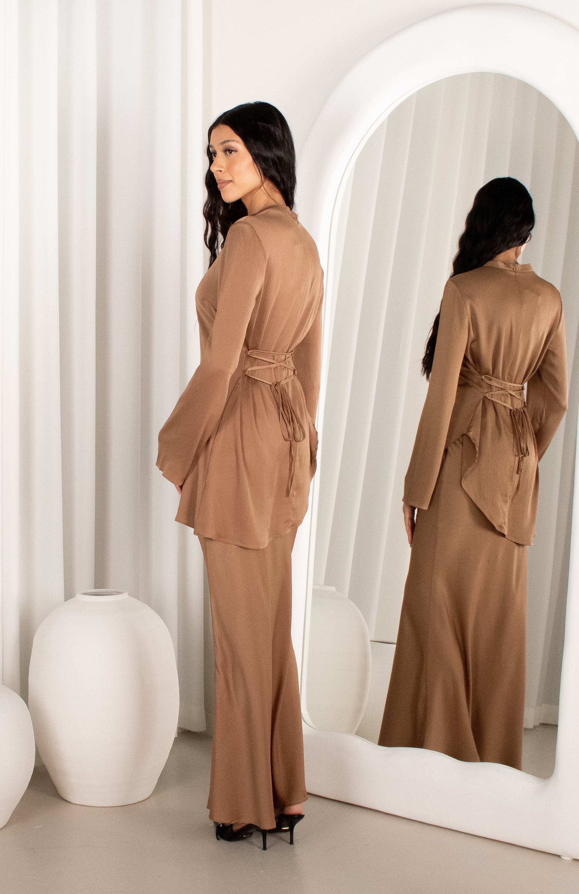 Woman in a brown outfit standing in front of a mirror in a minimalistic room.
