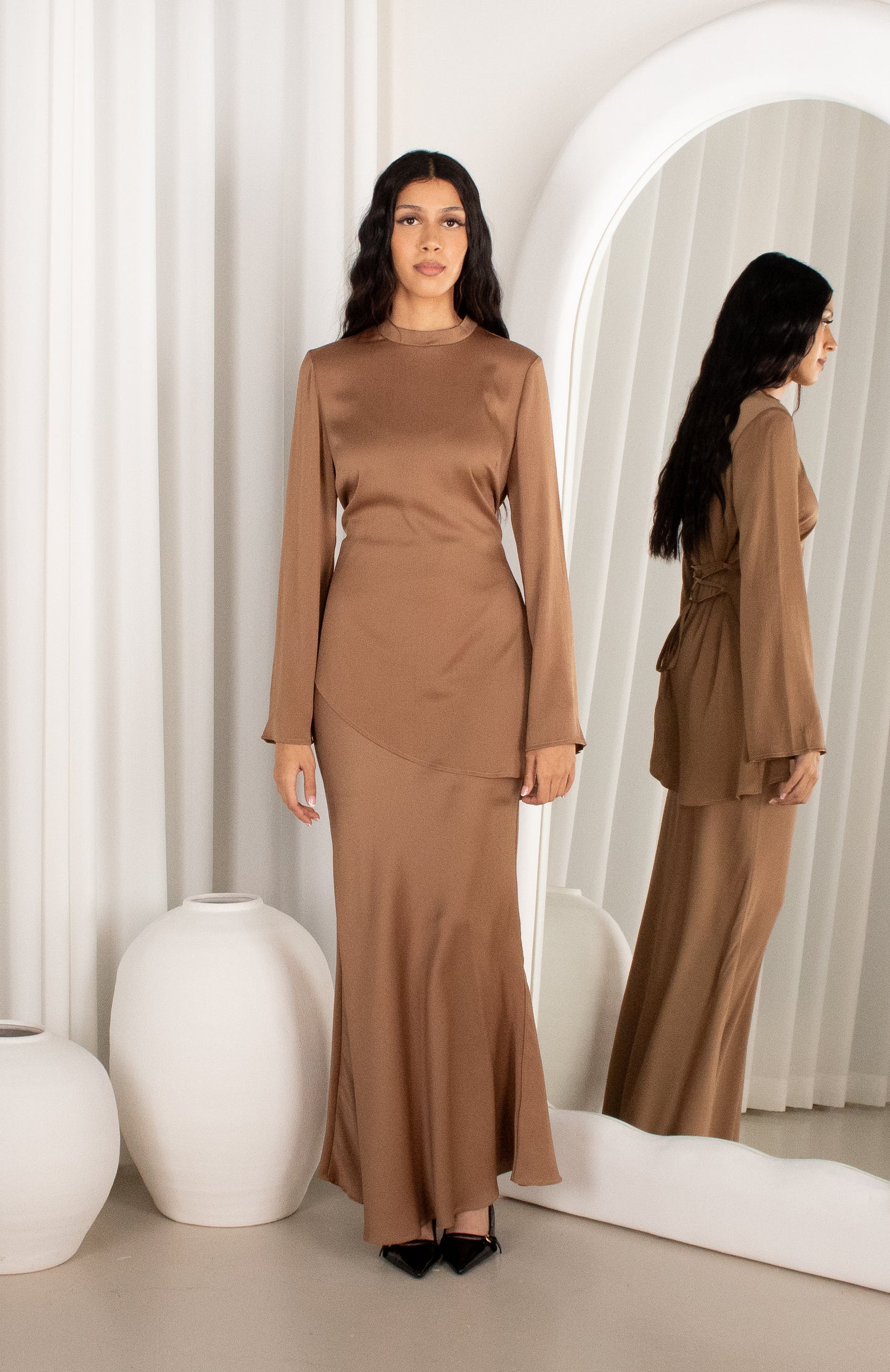 Woman wearing a brown outfit standing in front of a mirror with a white curtain background.