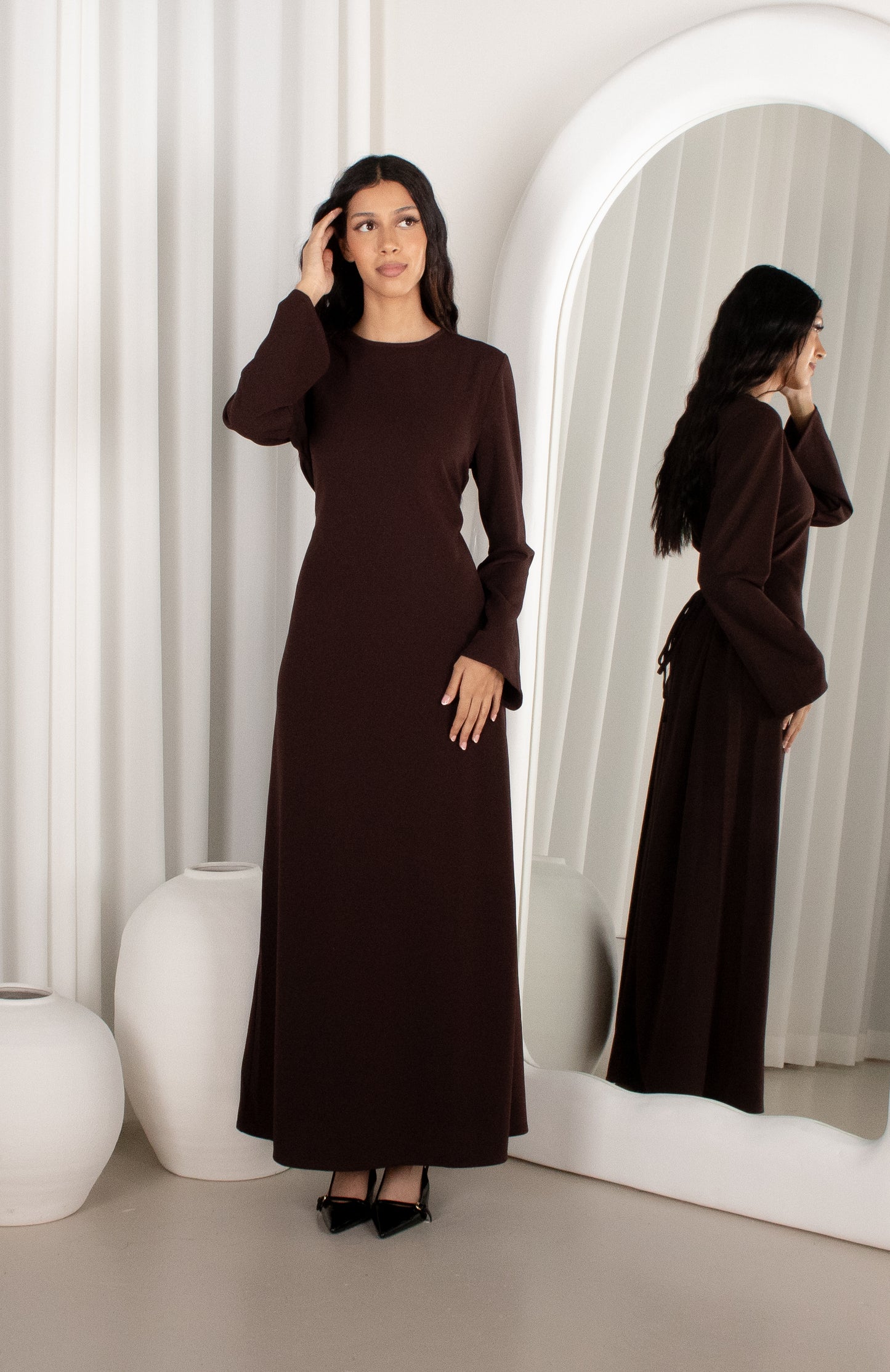 Woman in a brown dress standing in front of a mirror with white curtains and decor.