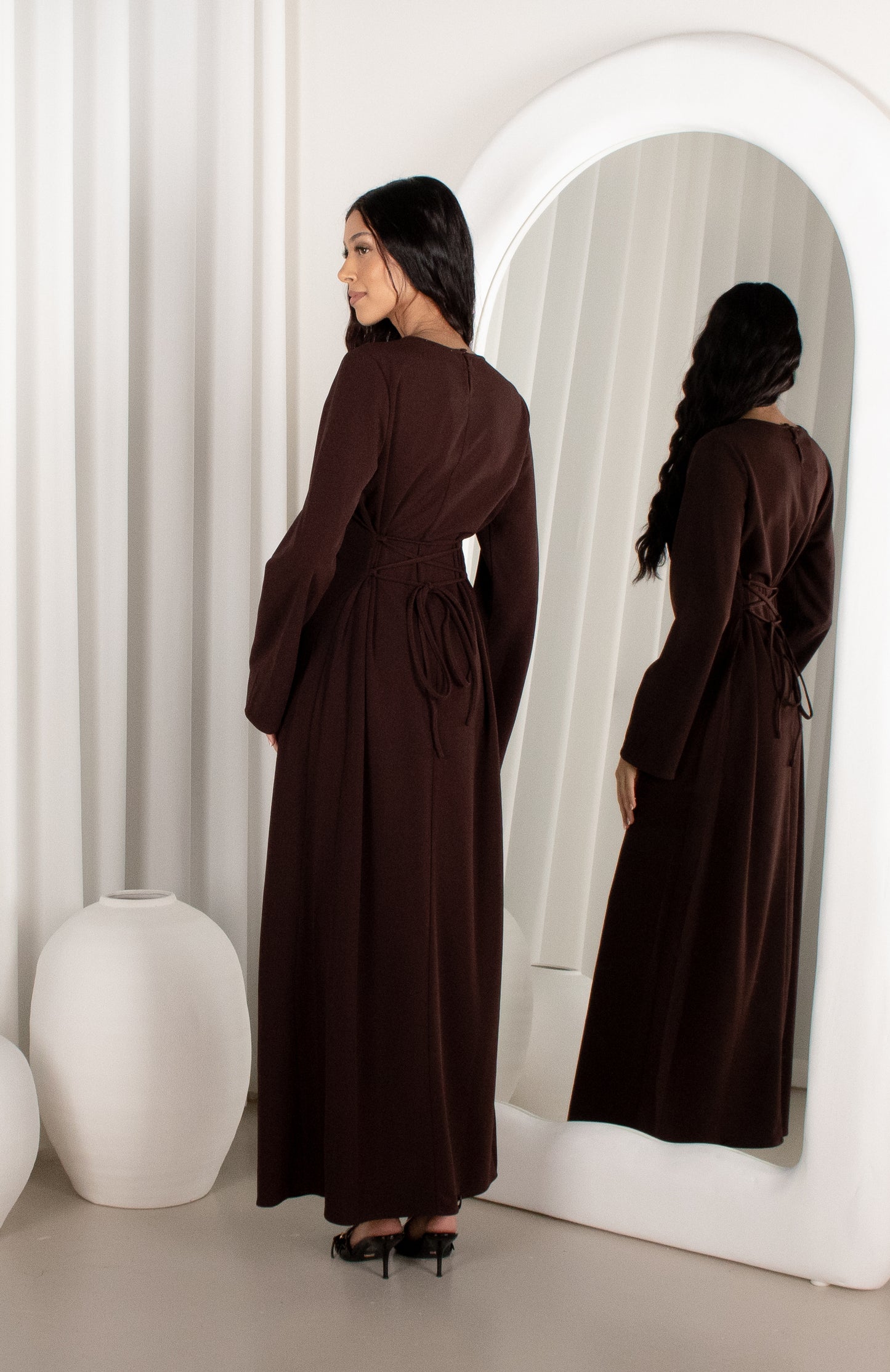 Woman in a brown dress standing in front of a mirror in a minimalistic room.
