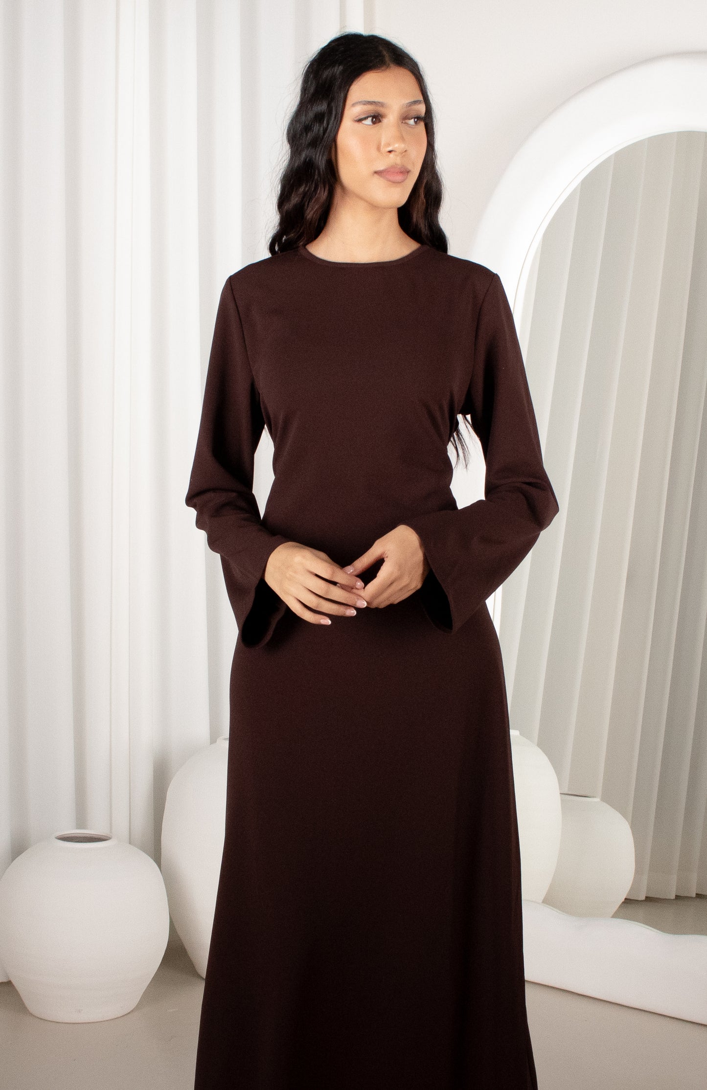 Woman wearing a brown dress in a minimalistic room with white curtains and decor.