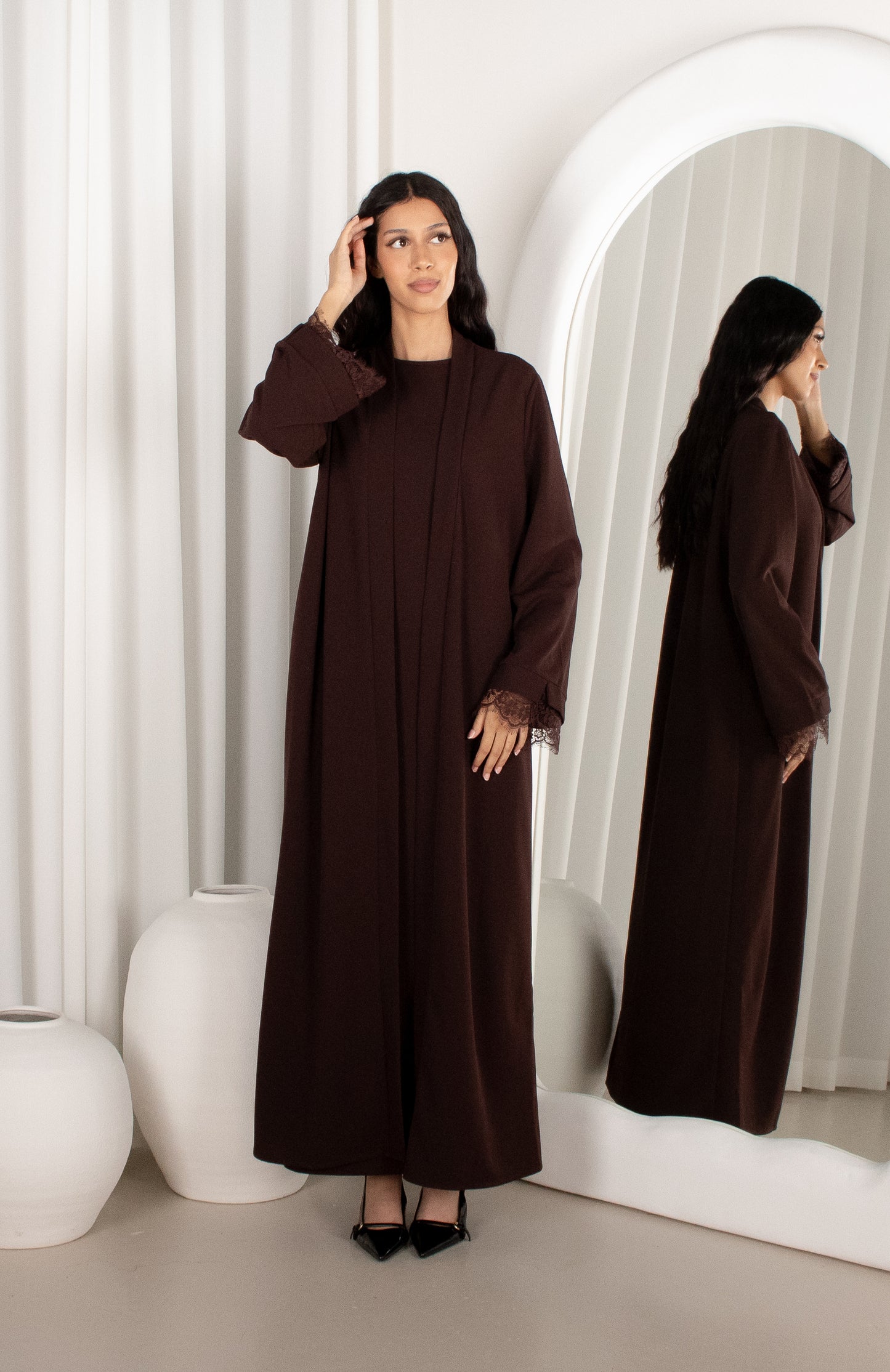 Woman in a brown abaya standing in front of a mirror in a minimalistic room.