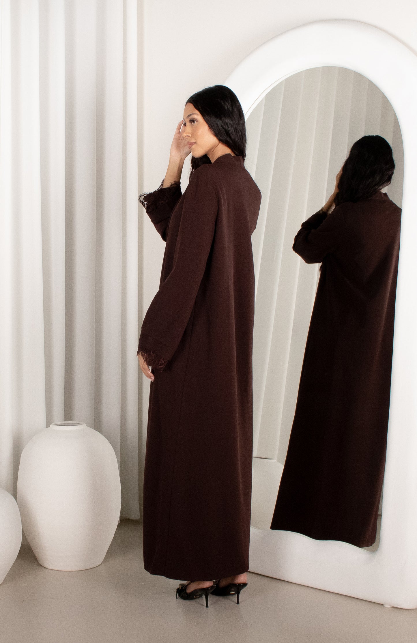 Woman in a brown abaya standing in front of a mirror in a minimalistic room.
