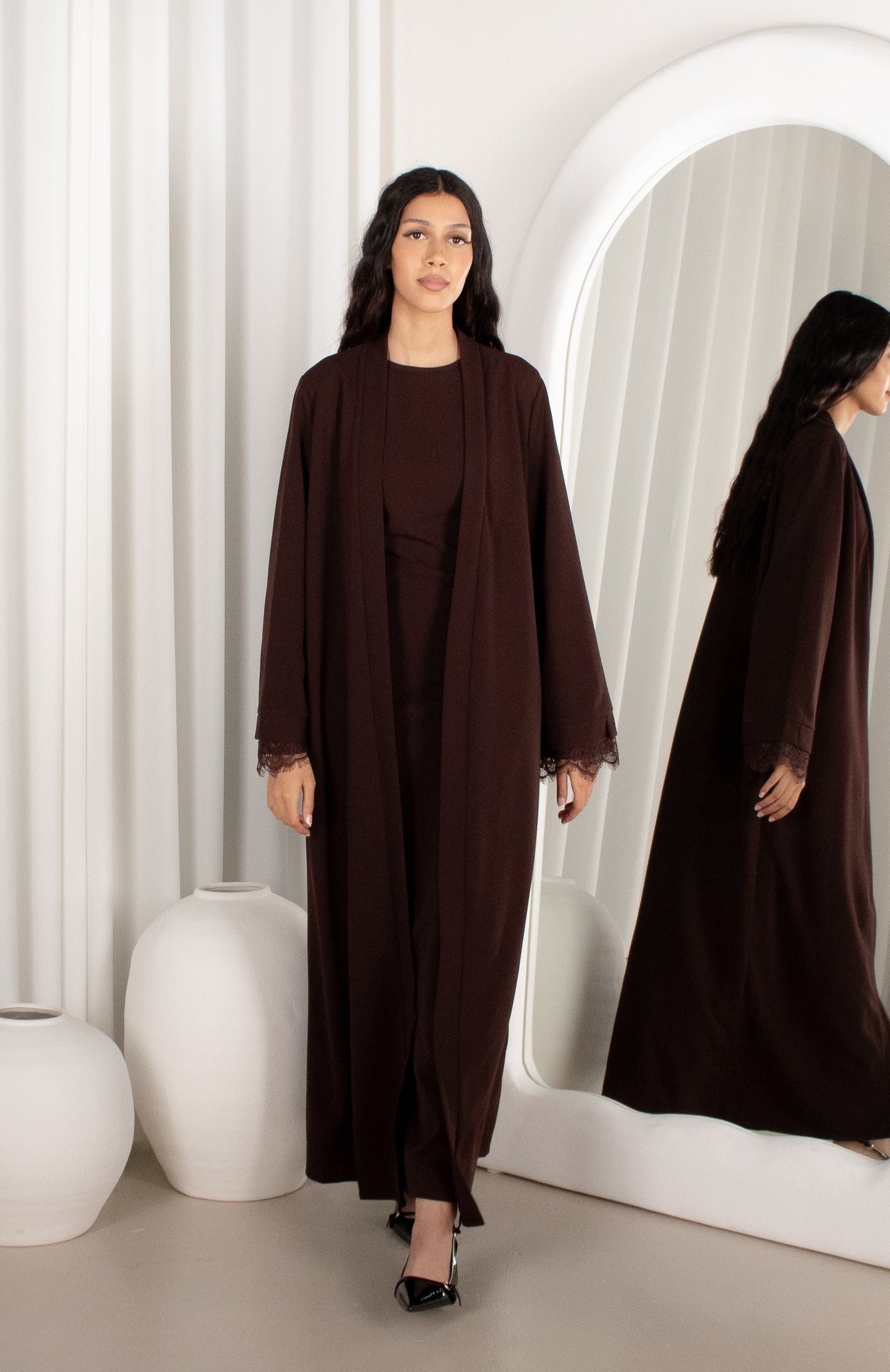 Woman in a brown abaya standing in front of a mirror with a white curtain background