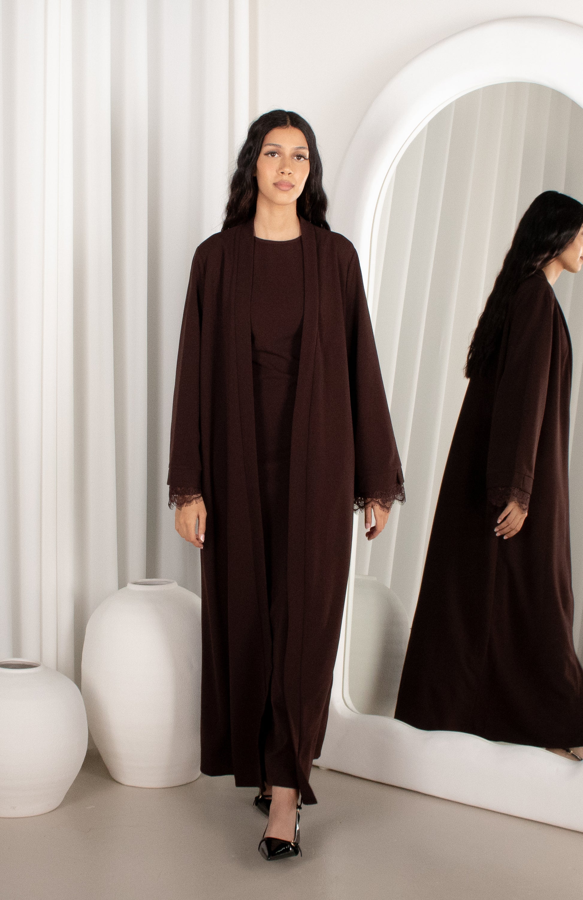 Woman in a brown abaya standing in front of a mirror with a white curtain background