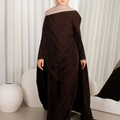 Woman wearing a brown off-shoulder dress with a draped design against a white curtain background.