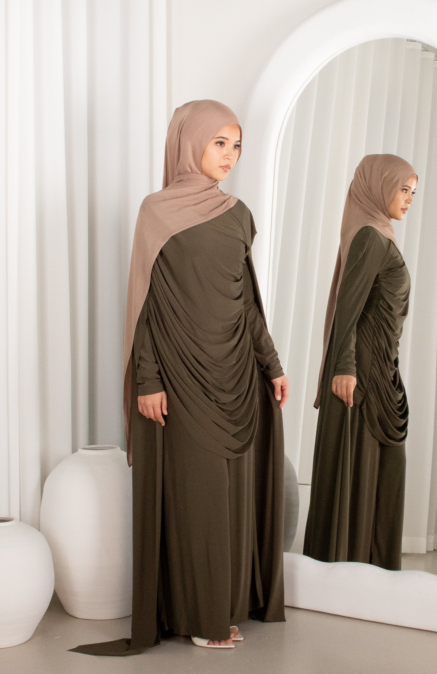 Woman in a long olive green dress with a matching hijab standing in front of a mirror.
