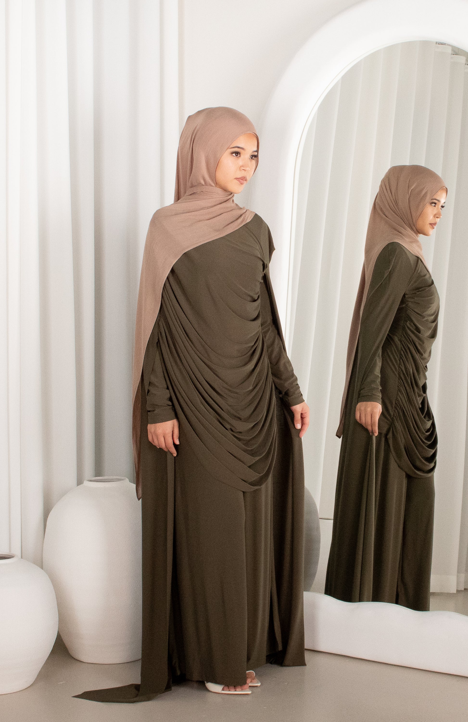 Woman in a long olive green dress with a matching hijab standing in front of a mirror.