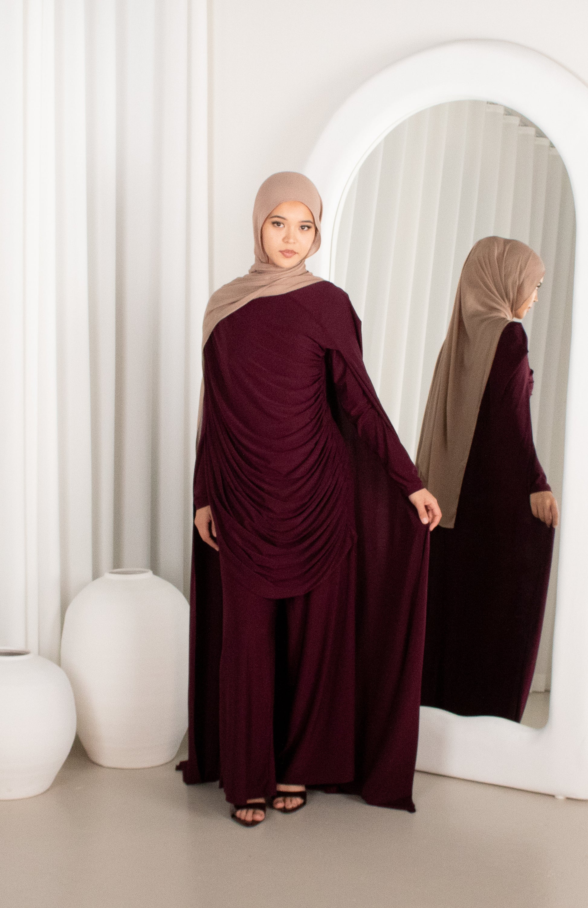 Woman in a burgundy dress and beige hijab standing in front of a mirror.