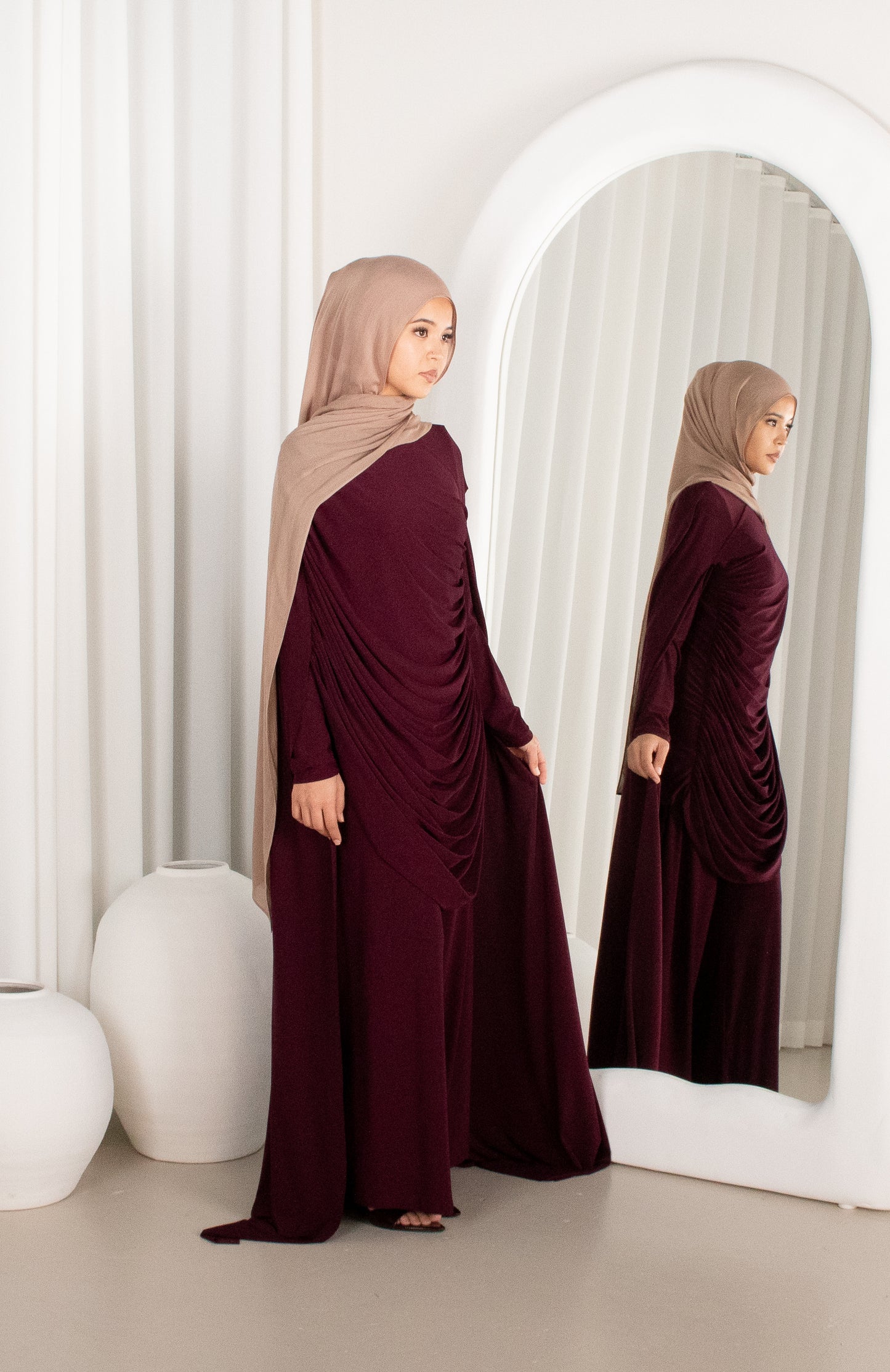 Woman in a burgundy dress and beige hijab standing in front of a mirror.