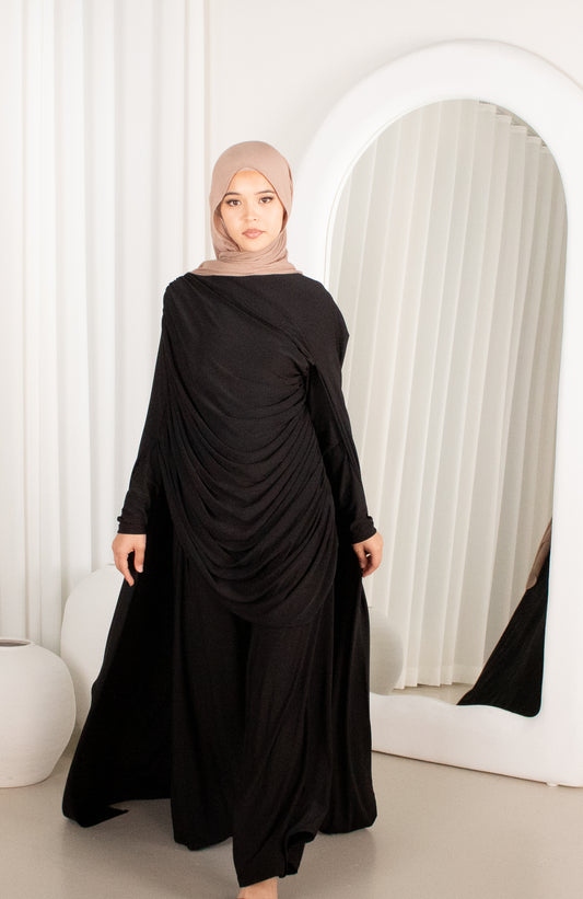 Woman wearing a black dress with a beige hijab in front of a mirror.