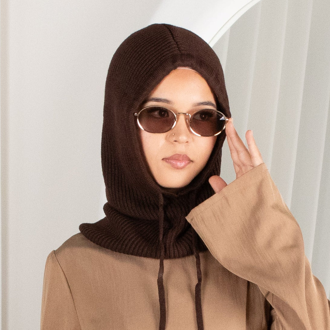 Woman wearing a brown outfit with a matching hijab against a white curtain background