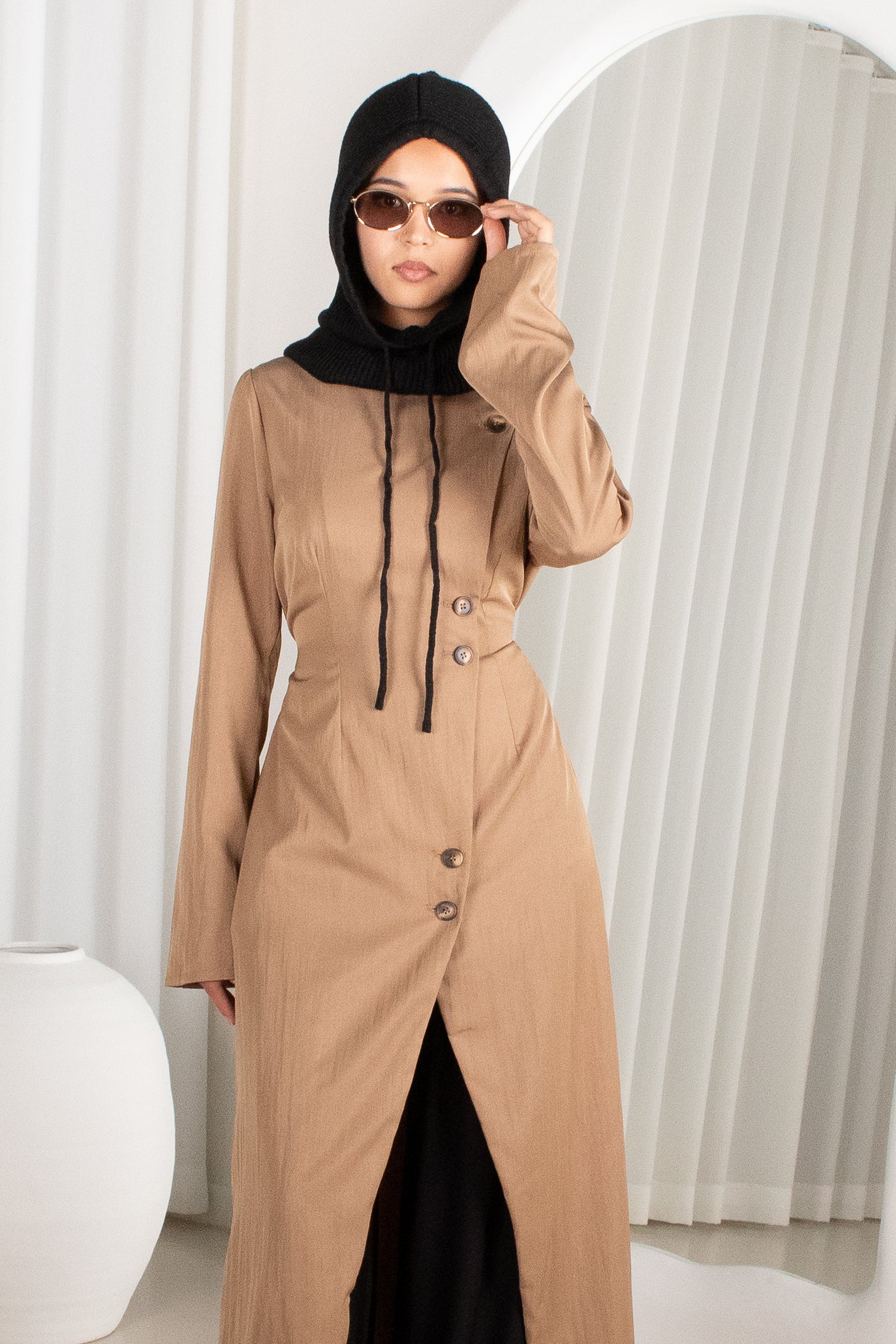 Woman wearing a beige long coat with black hijab and sunglasses indoors.