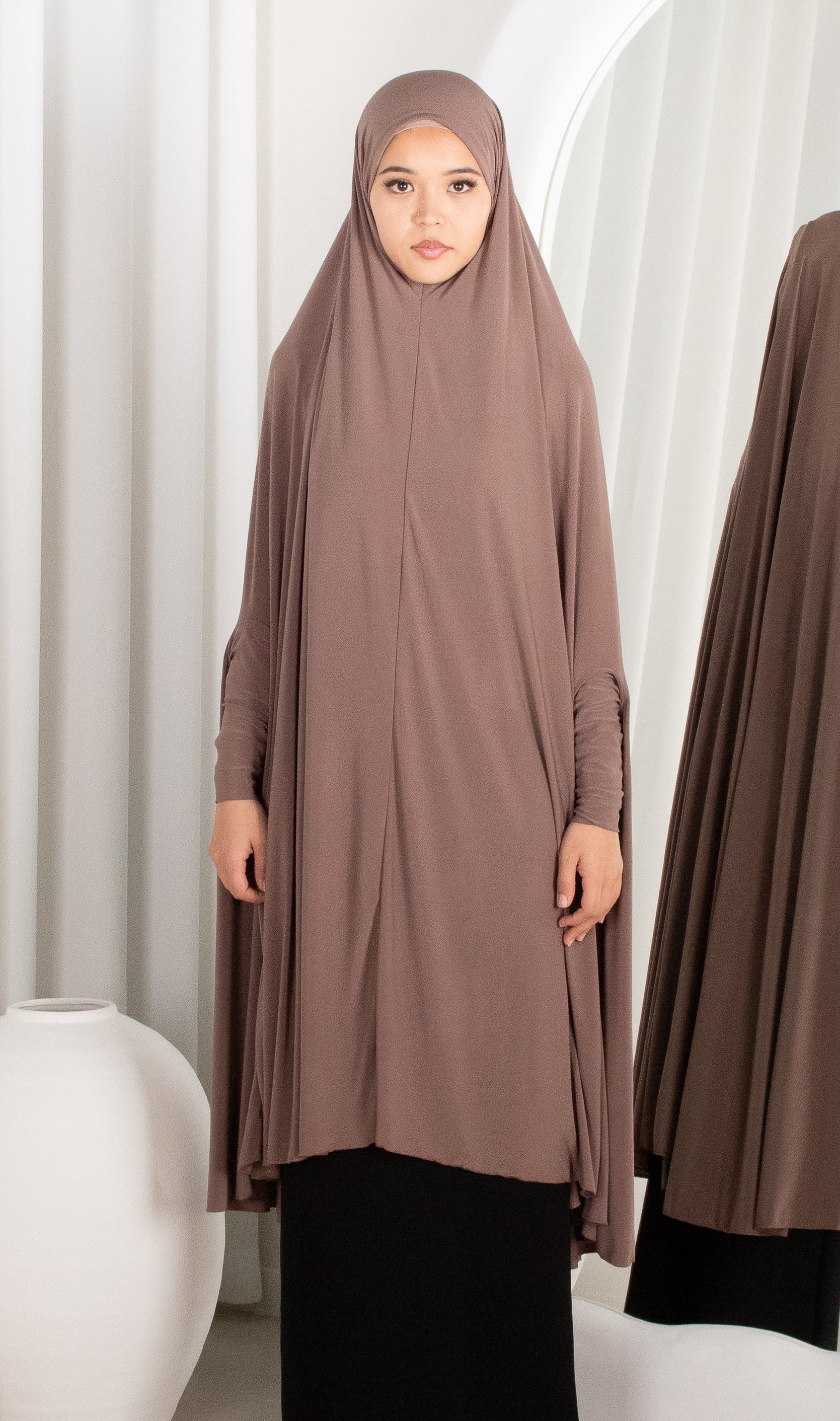 Woman wearing a brown hijab and dress in front of white curtains