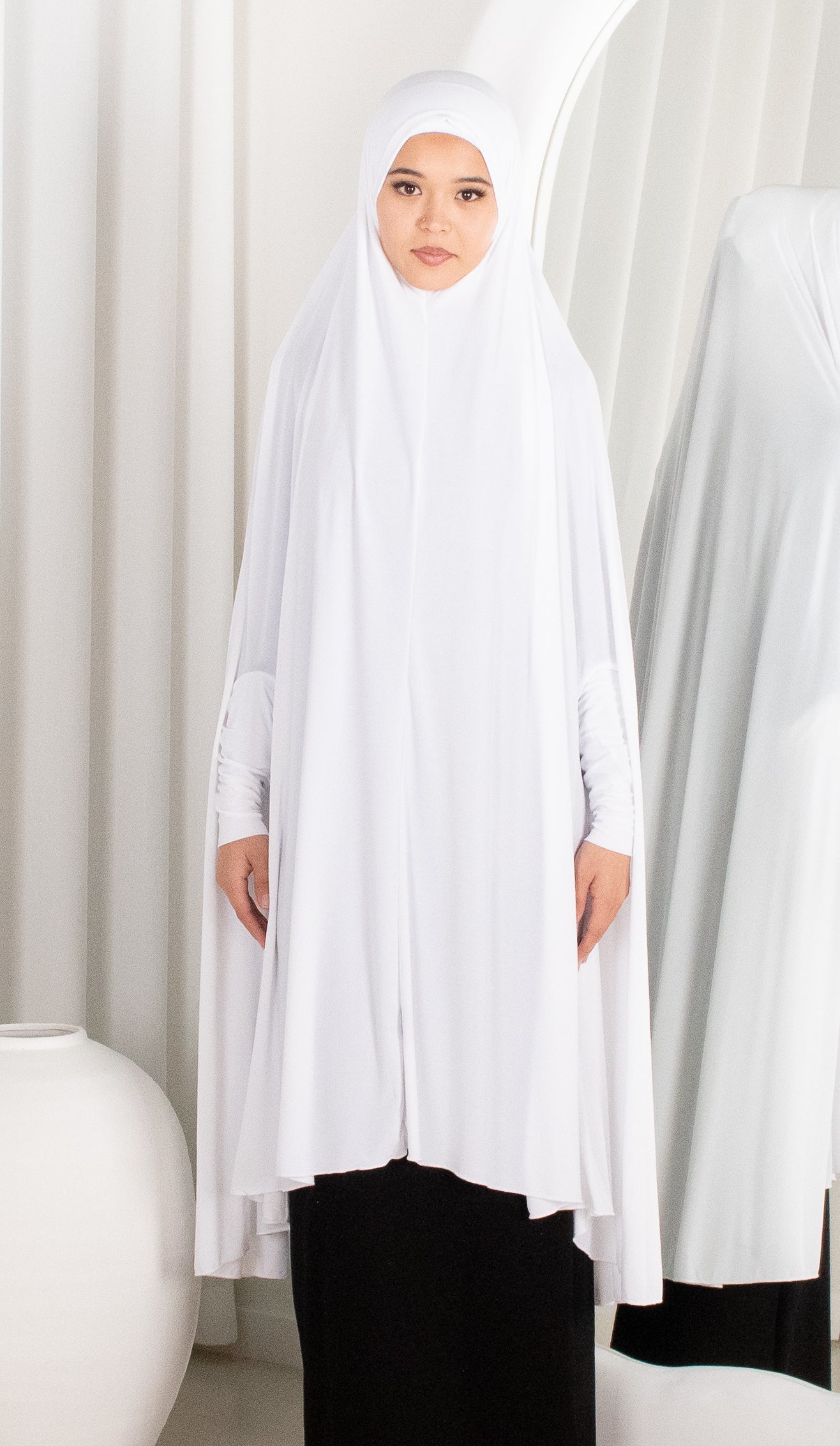 Woman wearing a white hijab and long white dress with black pants against a white curtain background