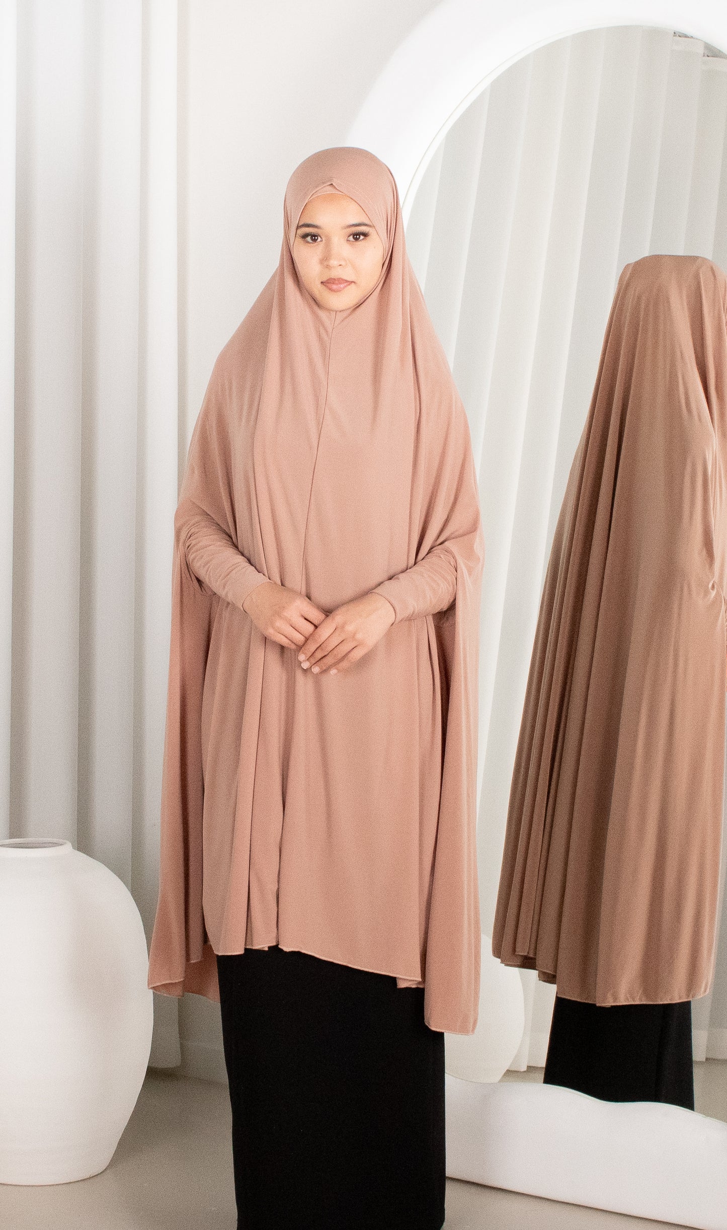 Woman wearing a beige hijab and black outfit in front of a mirror.