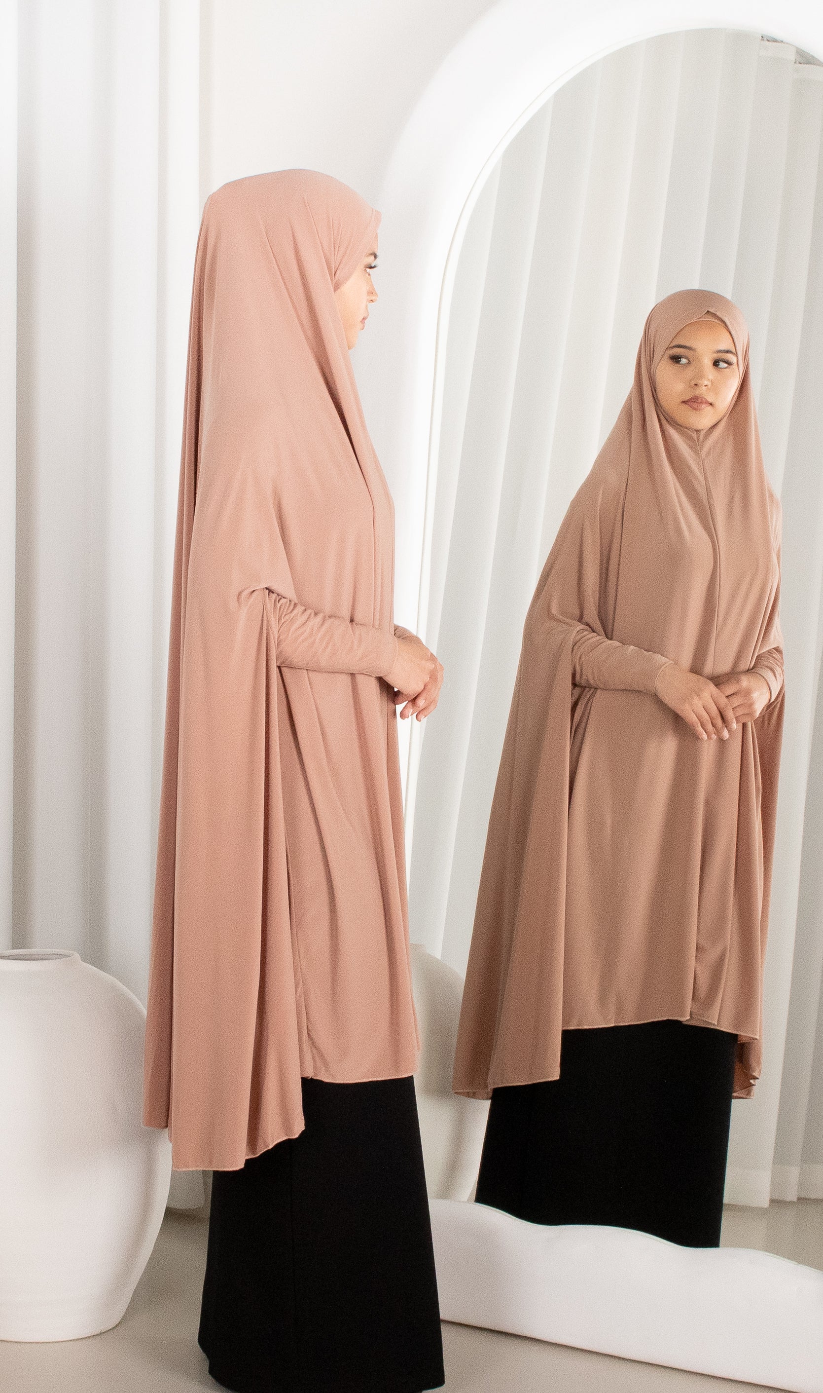 Woman wearing a beige hijab and black outfit standing in front of a mirror.