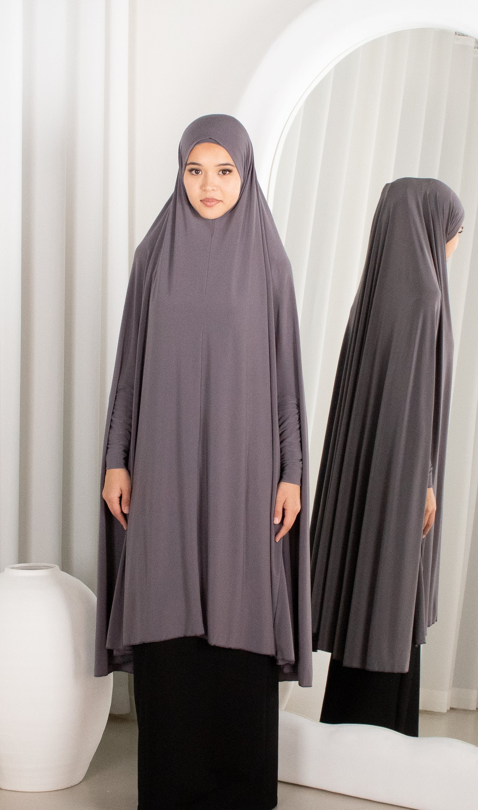 Woman wearing a gray hijab and dress standing in front of a mirror.