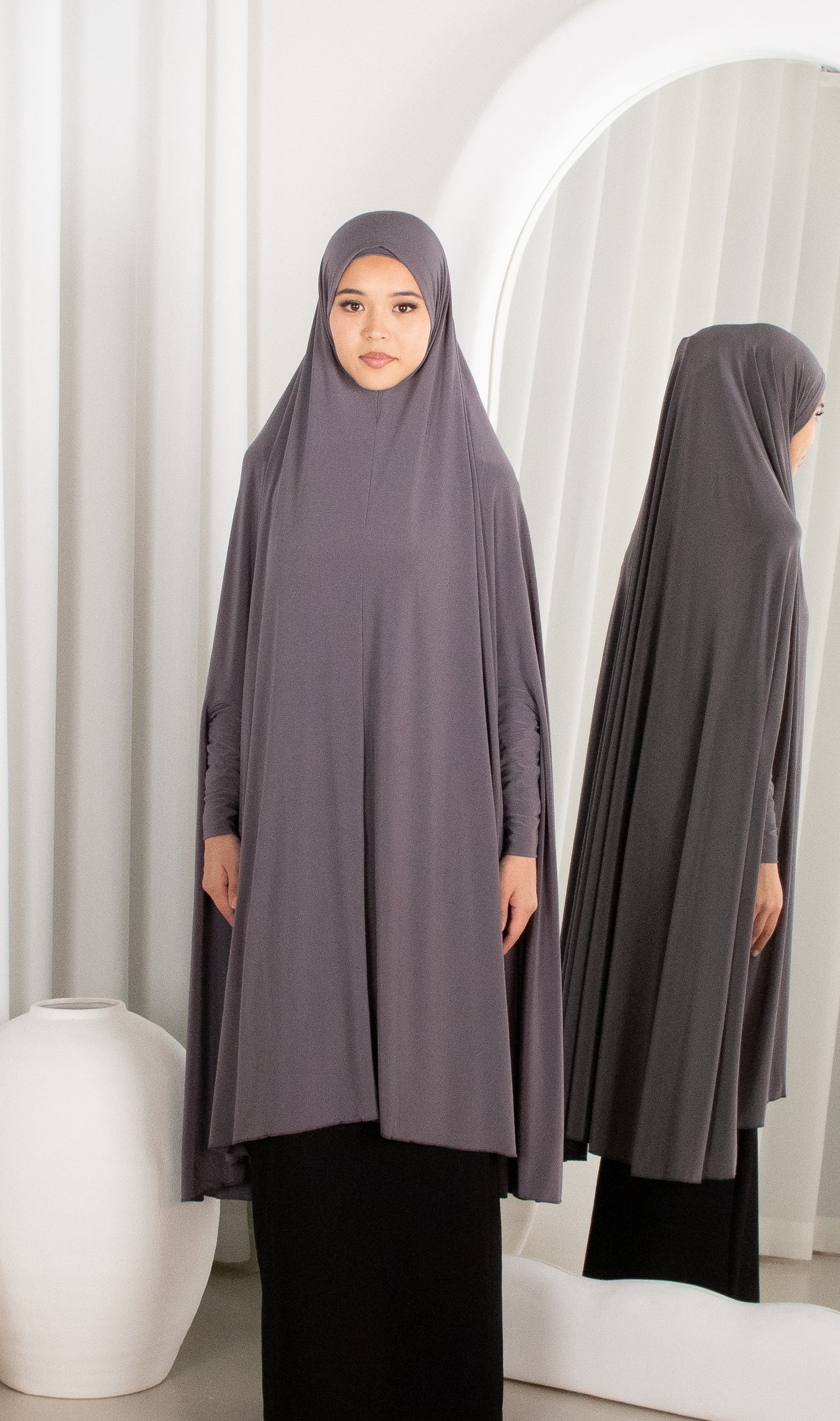 Woman wearing a long, light gray hijab in front of a mirror.